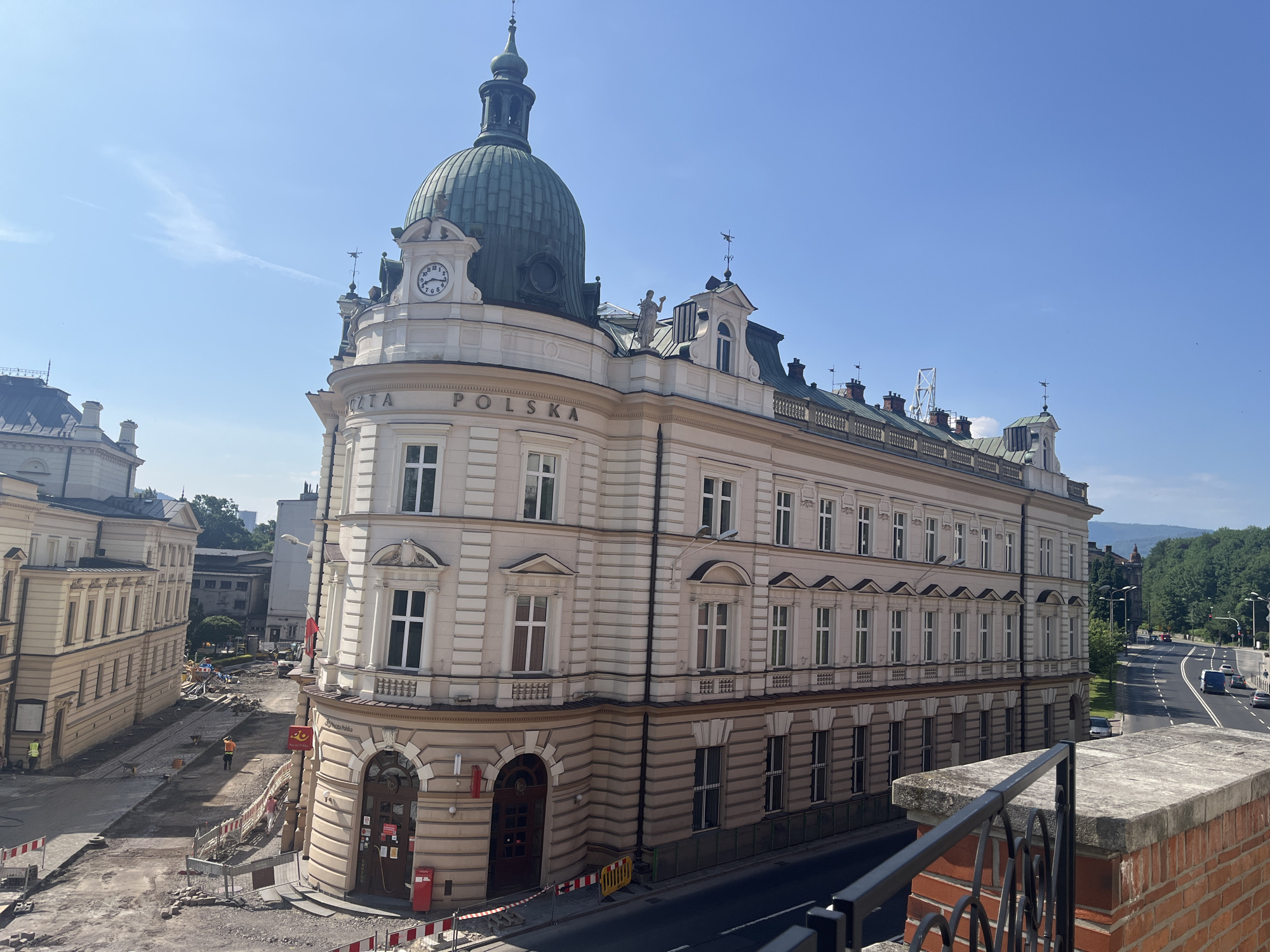 The image shows an exterior view of a cream-colored, multi-story building with a prominent green dome topped by a spire. The building features ornate architectural details, including arched windows, decorative moldings, and a clock face on one of its upper levels. The text "POCZTA POLSKA" is visible on the facade, indicating it is a post office.
In front of the building, there is a street undergoing construction, with visible barriers, piles of gravel, and construction equipment. A few construction workers can be seen in the area. To the left of the building, another similar-style building is partially visible. In the background, a road with traffic and lush green trees lining it can be seen, with hills or mountains in the far distance under a clear blue sky. The overall scene depicts an urban environment with ongoing civic work.
