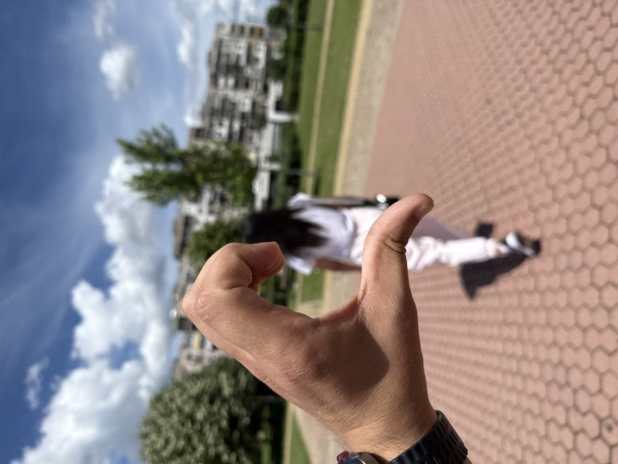 A close-up shot focuses on a human hand forming the left half of a heart shape in the foreground. The hand is tanned with visible skin texture and is positioned with the thumb and index finger extended, while the other fingers are curled inward. A dark-colored watch with a textured band is visible on the wrist.
In the background, a person is seen from behind, walking away from the camera. This individual appears to be wearing a white t-shirt and light-colored pants. Their dark hair is visible. The person's shadow is cast on the ground.
The environment is an outdoor urban setting. The ground in the foreground is paved with reddish-brown hexagonal tiles. Behind the paved area, there is a strip of green grass, followed by trees and in the distance, a multi-story building complex. The sky above is blue with scattered white, fluffy clouds. The lighting suggests it is daytime.
The scene is likely located in a park or public area within a city, possibly in Vratsa, Bulgaria, given the architectural style of the buildings and the general ambiance of a European city.