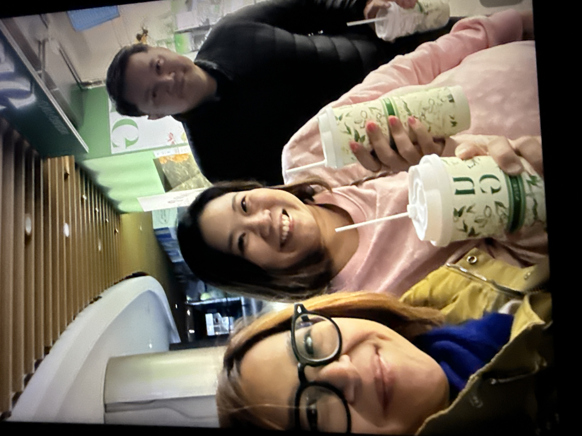 In Taipei, Taiwan, a group of three individuals, two women and one man, pose for a selfie in what appears to be an indoor public space with modern architecture. The woman in the foreground on the left wears eyeglasses and a mustard-yellow jacket over a blue scarf. To her right, another woman in a pink sweatshirt smiles warmly, holding a patterned green and white paper cup with a white lid and straw. To the right of these two women, a man in a black jacket smiles slightly, also holding a similar cup. The background features a bright green wall with white text and graphics, along with a wooden slatted ceiling. The overall atmosphere suggests a casual outing.