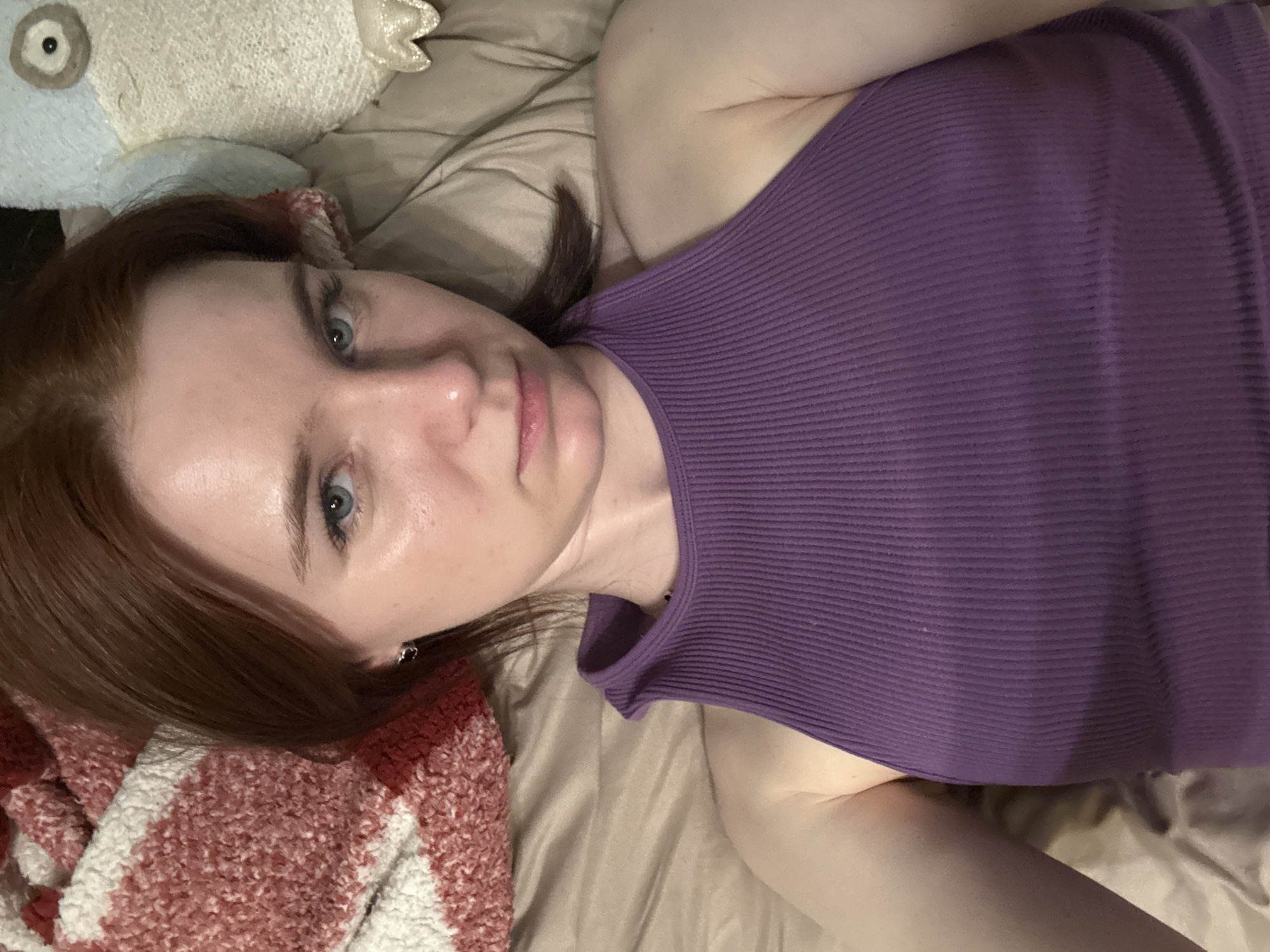 A pale-skinned woman with auburn hair and light-colored eyes is lying on a bed. She is wearing a purple, ribbed tank top. A stuffed animal is visible in the upper left corner of the image, and a fuzzy, patterned blanket is in the lower left corner. The bed has tan-colored sheets. The photograph appears to be taken from an overhead perspective. The location is unknown.