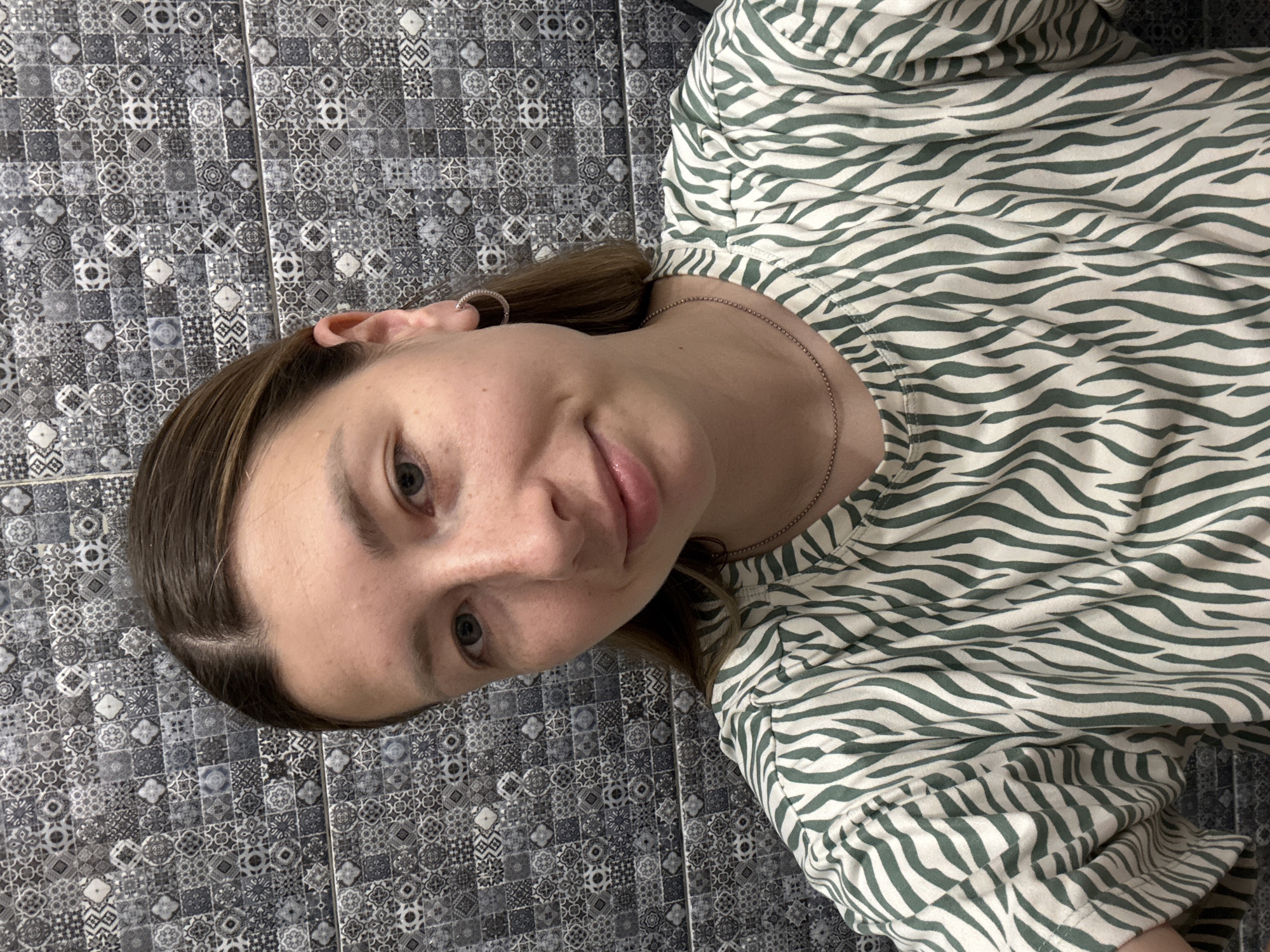 A young woman is taking a selfie in front of a tiled wall. She has light brown hair pulled back and is wearing a green and white striped shirt and a thin chain necklace with a small circular pendant. She has hoop earrings in both ears. The background is a wall covered in small, intricately patterned tiles, predominantly in shades of grey and black. The image appears to be taken indoors, possibly in a bathroom or kitchen, given the tiled wall. There are no visible indications of the specific location within Moscow, Russia.
