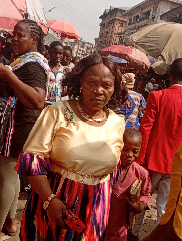 The image depicts a crowded outdoor market street in Onitsha, Nigeria, characterized by numerous individuals, market umbrellas, and multi-story buildings.
In the foreground, a woman with dark skin and dark, wavy hair looks directly at the camera. She wears a light gold long-sleeved top, a gold chain belt, and a long pleated skirt with vertical stripes of red, blue, purple, and black. A gold-colored choker necklace, a green and purple brooch, and a gold-colored bracelet are visible. She holds a small red clutch bag in her left hand.
To her right, a young boy with dark skin, wearing a maroon tunic, is partially visible. He holds a folded piece of gold-colored fabric or material in his right hand and looks towards the right, exhibiting a slight smile.
To the left of the main subject, another woman with braided hair is visible, wearing a black top, light leggings, and a multi-colored scarf draped over her left shoulder. She is looking towards the left.
The background features numerous individuals moving through the market. Overhead, several large market umbrellas in shades of red and beige provide shade. Behind the market structures, multi-story urban buildings with balconies are visible. The ground appears unpaved, suggesting a dirt or gravel surface. A person in the mid-right background wears a bright red jacket.