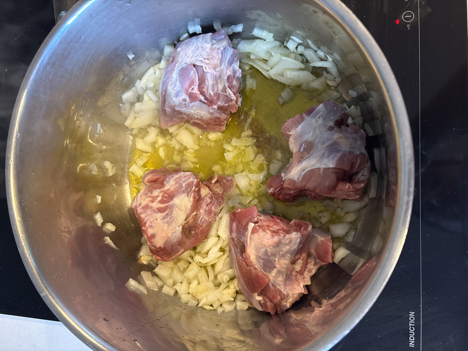 A stainless steel cooking pot is centrally positioned on a black induction stovetop. Within the pot, four distinct pieces of raw, bone-in meat, consistent with lamb or pork shoulder cuts, are visible. The meat is surrounded by and partially submerged in a quantity of yellow cooking oil and numerous diced white onion pieces. The stovetop surface to the lower left of the pot displays the white text "INDUCTION," and a small red indicator light is visible on the lower right. The immediate background is blurred. The scene depicts food preparation in a kitchen, located in Raanana, Israel.