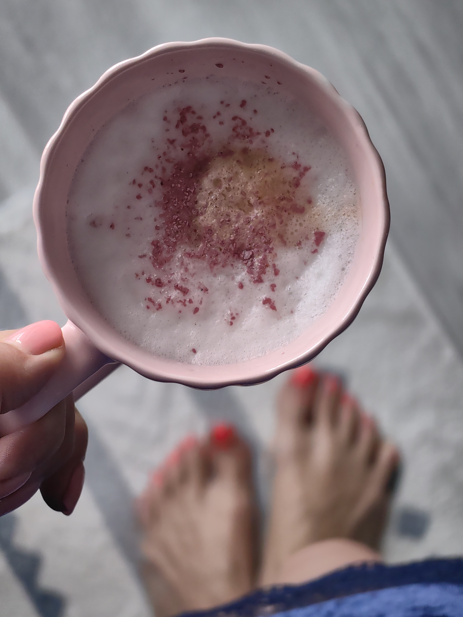 A close-up, overhead view shows a person holding a pink mug filled with a frothy, pink-tinged beverage. The mug has a scalloped edge and is positioned to the left of the frame. The beverage is topped with a layer of foam, sprinkled with small, reddish-pink pieces, possibly a spice or dried fruit. In the blurred background, two feet with bright red nail polish are visible on a light-colored surface. The overall lighting suggests a bright, possibly indoor environment with soft shadows. The location in Fanipal, Belarus, is not evident from the image's visual content.