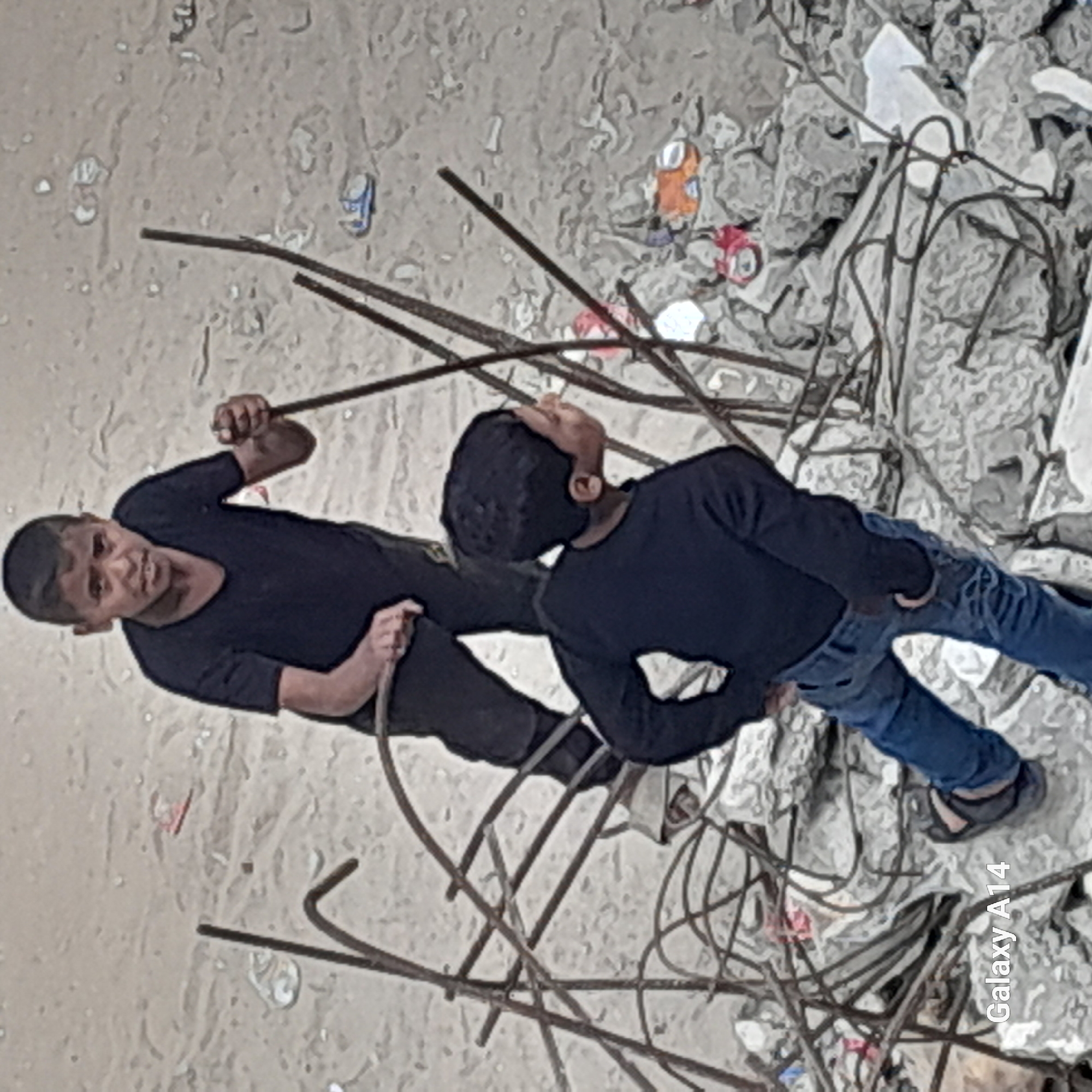 Two boys stand amid rubble and twisted metal rods. One boy, wearing a black shirt and dark pants, holds a rod and looks directly at the camera. The other boy, dressed in a black shirt and blue jeans, looks upward. The ground is a mix of dirt and debris, including discarded cans. This scene is in the city of Unknown, Unknown.