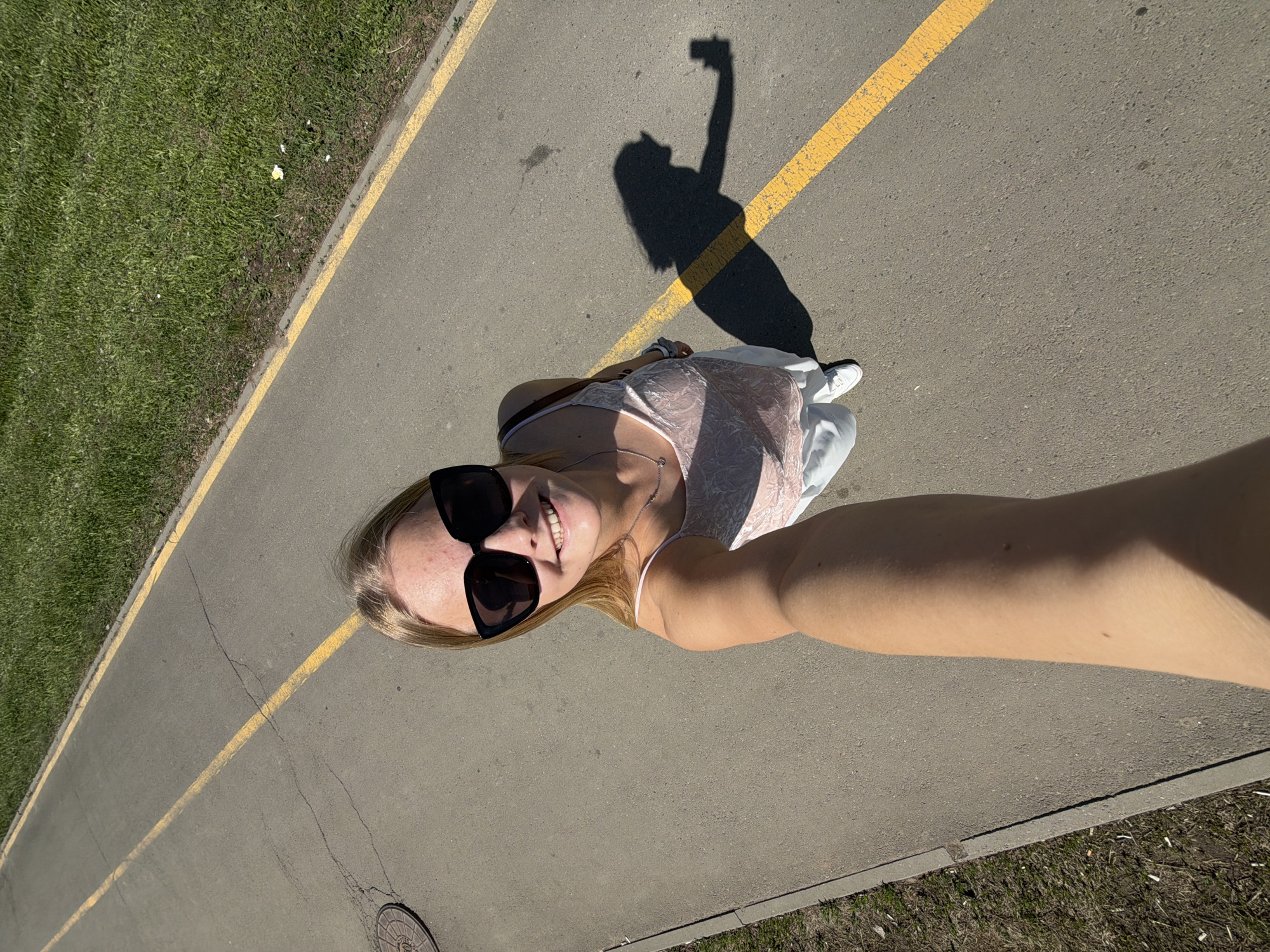 A fair-skinned, blond woman wearing sunglasses is taking a selfie on an asphalt walkway marked with a yellow line. She is wearing a light-colored top and pants, and white sneakers. Her shadow, holding a phone, is cast on the walkway next to her. To the left of the walkway is a grassy area. To the right is another area of ground cover and a sewer grate. The location is Izhevsk, Russia.