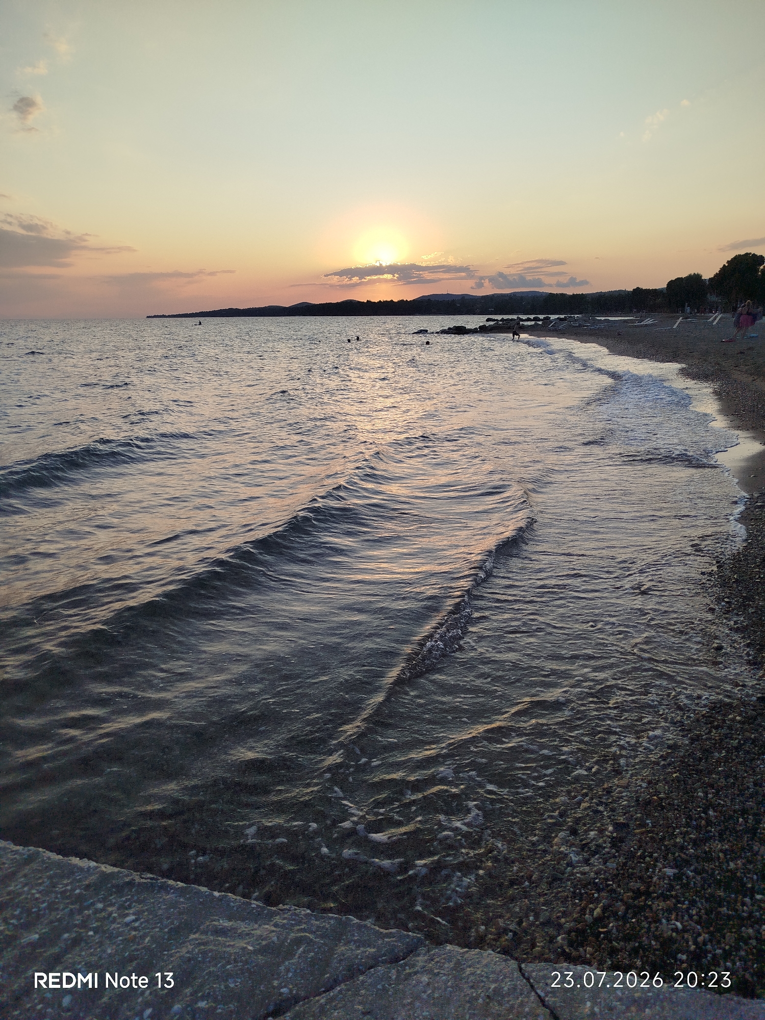 A sunset scene unfolds over the Aegean Sea, as viewed from the shoreline in Sithonia, Greece. The sun, partially obscured by clouds, casts a warm, golden glow across the sky and reflects on the gentle waves. The water laps at a pebble beach, where a few people can be seen in the distance enjoying the evening. The foreground features a concrete embankment, worn by time and the sea. The overall atmosphere is serene, capturing a tranquil moment at dusk.