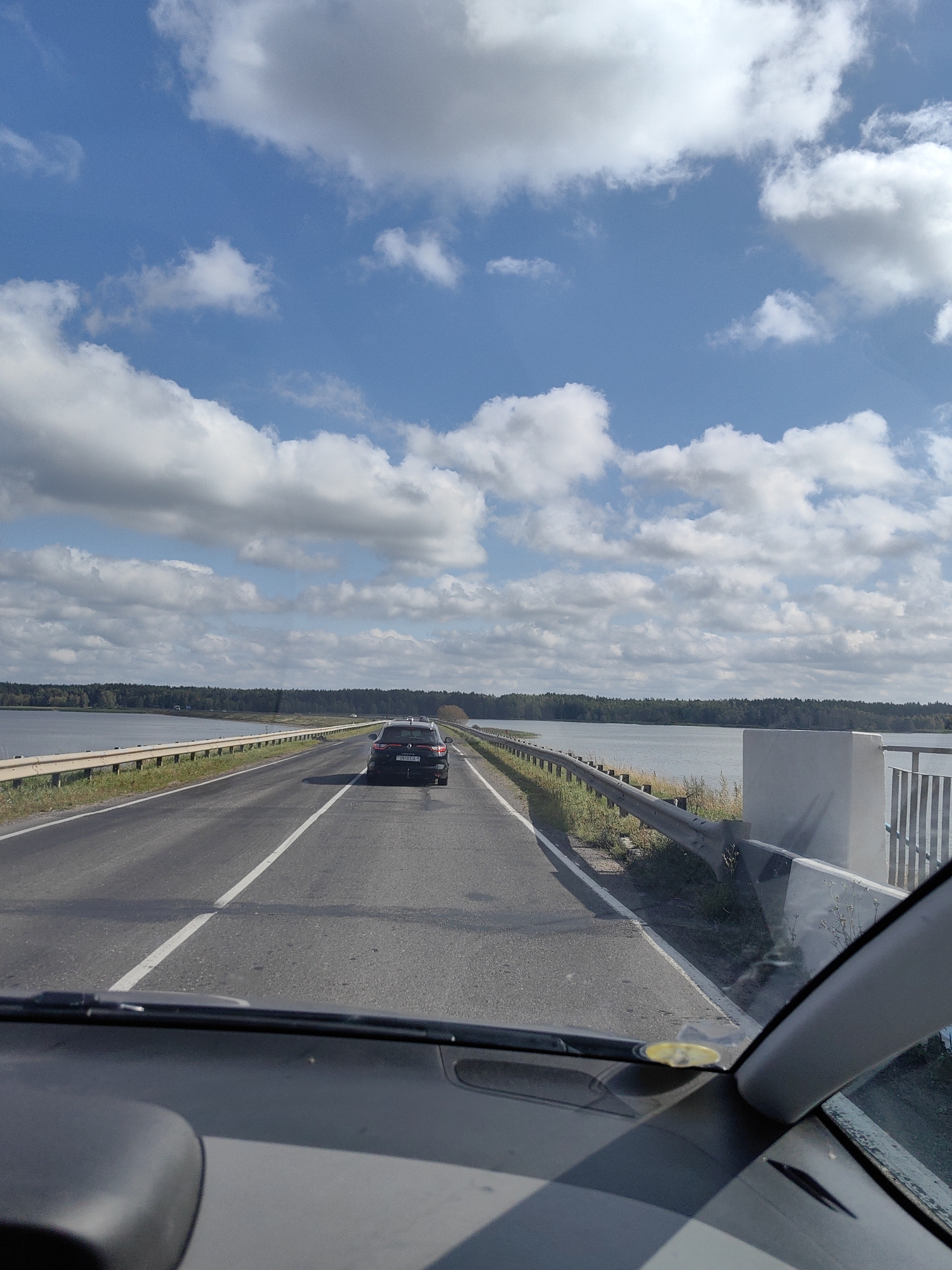 The image shows a view from inside a car traveling on a road that appears to be a bridge or causeway over a body of water. The road is paved, with a white dashed line indicating a lane separation. A dark-colored SUV is visible ahead on the road, also traveling in the same direction. The vehicle is positioned in the right lane.
On either side of the road, there is a metal guardrail. Beyond the guardrail to the left, a large body of water, likely a lake or river, stretches out, bordered by a dense line of trees. To the right of the road, the guardrail continues, and a portion of a concrete structure is visible.
The sky is blue with scattered white and gray clouds, suggesting a partly cloudy day. The overall environment appears to be rural or semi-rural, with no other significant structures or human activity visible in the immediate vicinity. The perspective of the image is from the driver's seat, looking forward through the windshield. The dashboard of the car is partially visible in the foreground. The location is identified as Kryvaselski Sel'ski Savet, Belarus.