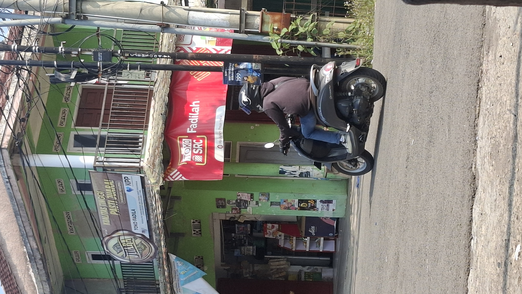 The image depicts a street scene in Cilaku, Indonesia. A person wearing a dark hoodie, blue jeans, and a helmet rides a dark-colored scooter on an asphalt road. To the left of the road is a two-story building with a light green facade. A banner with the text "KUALITAS TINGGI PASTI JUARA!" is prominently displayed on the upper level of the building. Below this, a red banner with "TOKO SRC Fadilah" is visible above the entrance to a shop. Several electrical poles and wires are present on the right side of the image, along with overgrown vegetation. The building appears to be a shop or a residence with commercial activity. The lighting suggests it is daytime with bright sunlight.