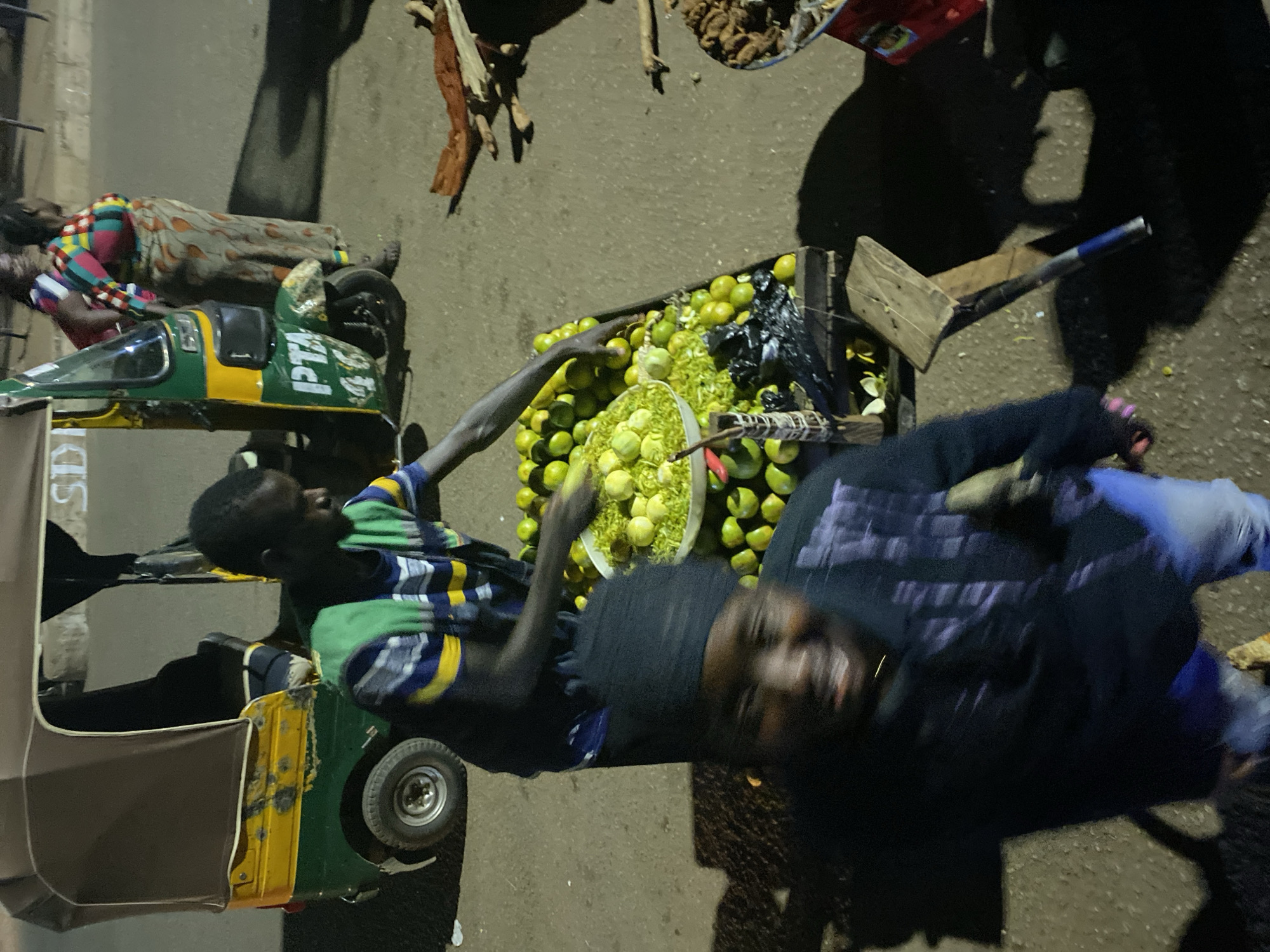The scene is a street in Bukuru, Nigeria. A man wearing a blue and yellow striped shirt is selecting lemons from a crate. A second man in a dark hooded shirt is looking towards the camera. The crate of lemons is next to a green and yellow auto rickshaw. Another person, dressed in multi-colored clothing, is lying down near the auto rickshaw. There is a container with dark, unidentifiable food items. The environment is urban, dimly lit, and appears to be either early morning or late evening.