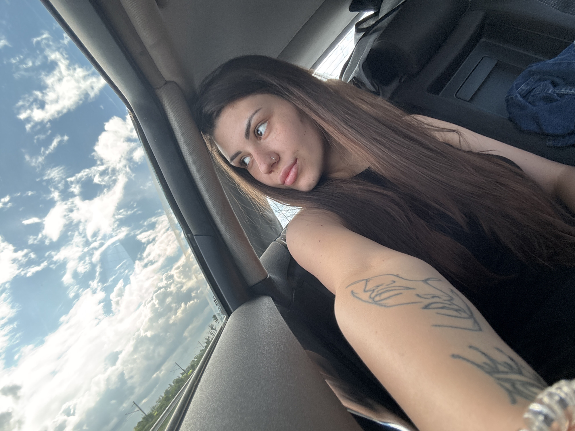 A young woman with long, straight dark brown hair is captured from the chest up, leaning against a vehicle's window frame. She has a calm, slightly pensive expression, with her gaze directed outside the window. She has visible freckles across her nose and cheeks and wears a silver nose stud. On her left arm, two tattoos are visible: an abstract, stylized face or figure on her upper arm, and a less distinct design, possibly geometric, further down her forearm. She is also wearing a beaded or crystal bracelet on her left wrist and a dark, likely black, sleeveless top.
The setting appears to be the interior of a moving car or bus, with parts of the vehicle's dark gray door frame, a headrest, and storage compartments visible. Through the window, a bright blue sky dominates the view, filled with scattered, fluffy white clouds. Below the sky, a hint of a landscape with green fields or trees and utility poles can be seen, suggesting a rural or suburban journey.
The scene is illuminated by bright daylight, indicating late morning or afternoon under fair weather conditions. The overall atmosphere is tranquil. The location context provided is Herakovo, Bulgaria. There is no visible text in the image.