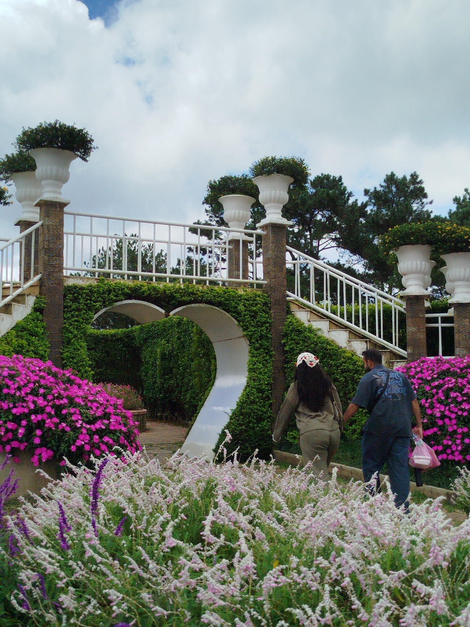 The image captures a couple walking through a lush, landscaped garden, likely in the city of Lâm Viên - Đà Lạt, Vietnam, known for its picturesque scenery. In the foreground, a profusion of delicate pale pink and purple flowers creates a soft, textured carpet, interspersed with taller stalks of purple blooms. Behind this floral display, vibrant pink flowers in a large planter overflow to the left.
A white ornamental railing runs along the top of a stone and brick structure that supports a wide staircase. This staircase ascends to the right, flanked by ornate white urns topped with green foliage. The structure is enveloped by dense, manicured green hedges, forming elegant arches.
The couple walks away from the camera, towards the staircase. The woman, on the left, has dark hair and wears a muted olive-green outfit, with a white and red floral headscarf. The man, on the right, is dressed in a dark blue t-shirt and dark pants, carrying a pink plastic bag in his right hand. They appear to be walking together.
The background is dominated by tall, dark green pine trees reaching towards a cloudy sky. The overall atmosphere is serene and natural, suggesting a leisurely stroll in a well-maintained botanical garden or park.