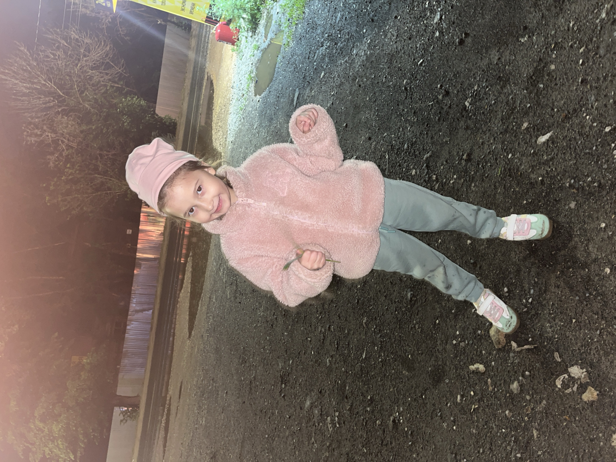 A young girl stands outdoors on a dark surface, possibly pavement or asphalt, with scattered debris. She wears a pink fuzzy jacket, light green pants, and colorful sneakers with white straps. A pink hat sits on her head. She holds a small green plant in her hand. The background shows trees and a fence, illuminated by artificial light that suggests it's evening or night. Puddles are visible, indicating recent wet conditions. The image's orientation is tilted. Novokuybyshevsk, Russia cannot be determined from the visual information alone.