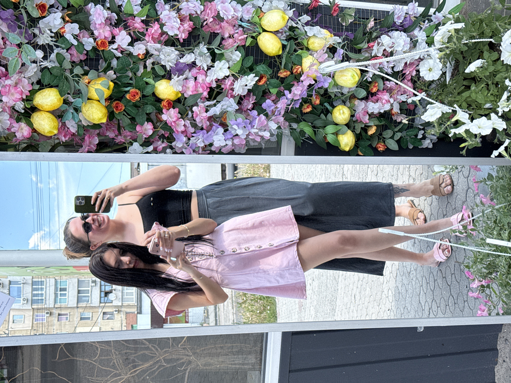Two young women are reflected in a large mirror outdoors. The woman on the left, with long dark hair, is wearing a light pink short dress with buttons down the front and is holding a pink phone. The woman on the right, with her hair in a bun and wearing sunglasses, is wearing a black tank top and a dark grey skirt, and is holding a phone. Behind them, a wall is decorated with an artificial floral arrangement featuring pink, purple, and white flowers, green leaves, and yellow lemons. The ground is paved with gray interlocking bricks. Buildings and a cloudy sky are visible in the reflection of the mirror.