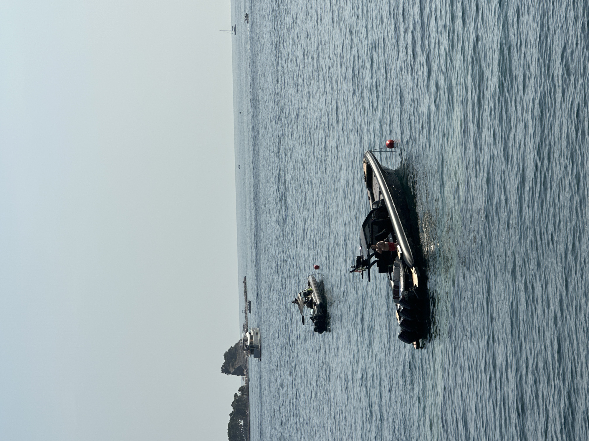 An aerial view captures the serene waters off the coast of Budva, Montenegro, where several vessels navigate the calm sea under a clear, pale sky. The foreground is dominated by a sleek, black rigid inflatable boat (RIB) with multiple outboard engines, its sleek design hinting at speed and power. A lone individual, clad in red shorts, stands on the deck, appearing to observe the surroundings. A red buoy floats nearby, tethered to the RIB by a thin rope, a common marker in maritime settings.
Further out in the middle ground, another RIB, this one a lighter shade, is moored. Two individuals are seated within its protective canopy, their presence suggesting a moment of rest or observation. In the distance, a larger yacht cruises on the horizon, its form softened by the atmospheric haze. Behind the yacht, a rocky outcrop with scattered vegetation rises from the sea, characteristic of the Budva coastline. Several smaller boats are also visible, dotting the expanse of water, contributing to the sense of a tranquil maritime activity. The overall scene conveys a sense of leisure and exploration on the Adriatic Sea.