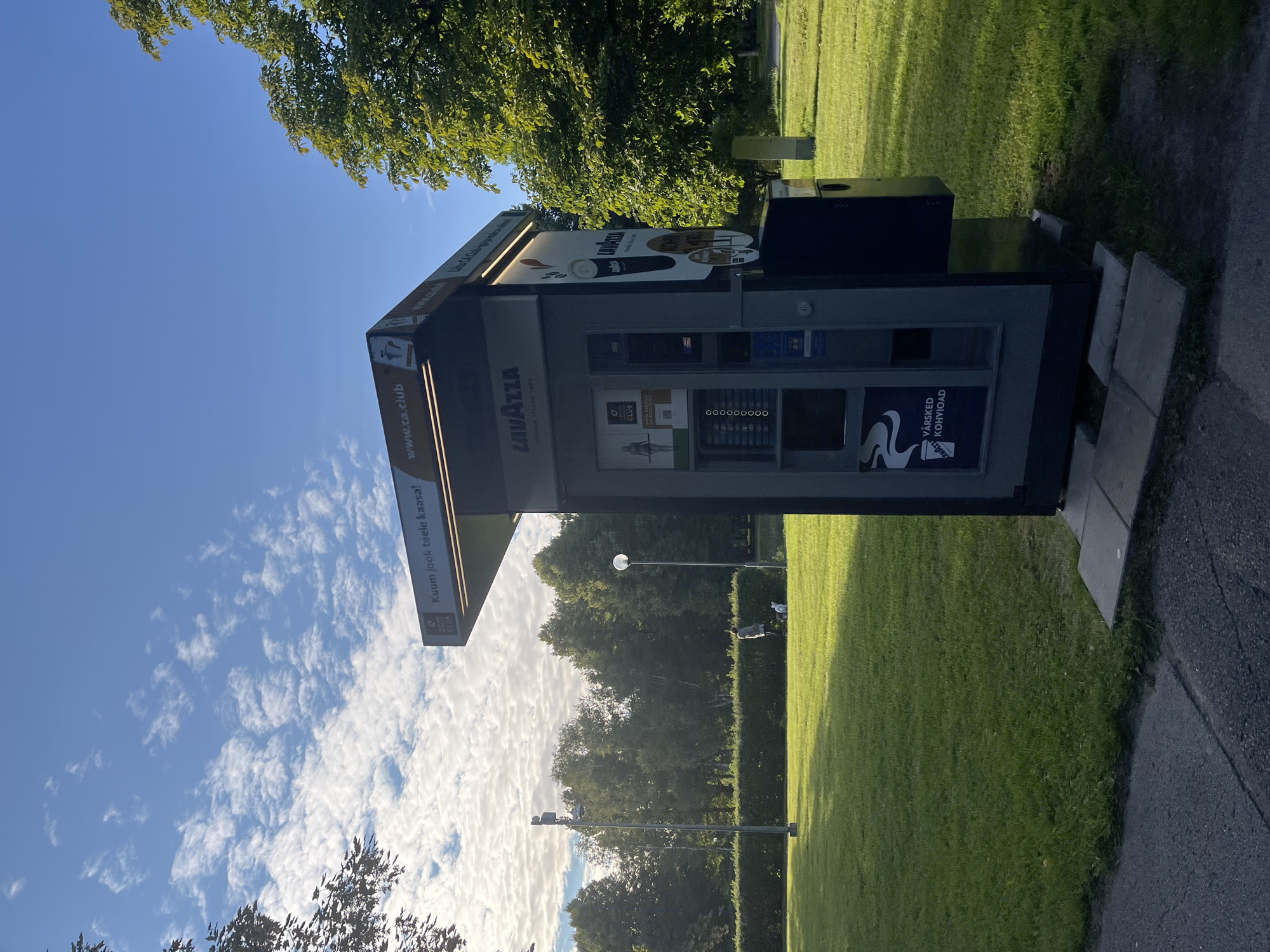 A Lavazza coffee vending machine is situated outdoors on a sunny day, with a person and two dogs visible in the background on a green lawn. The vending machine is grey and black, with the Lavazza logo and branding prominently displayed. It has a digital display screen and slots for cups and payments. The top of the machine features a sign with the text "Kuumi jook teele kaasa!" and "www.ca.club". The environment includes a grassy area, trees, a sidewalk, and a clear blue sky with scattered clouds. The overall impression is of an urban park setting.