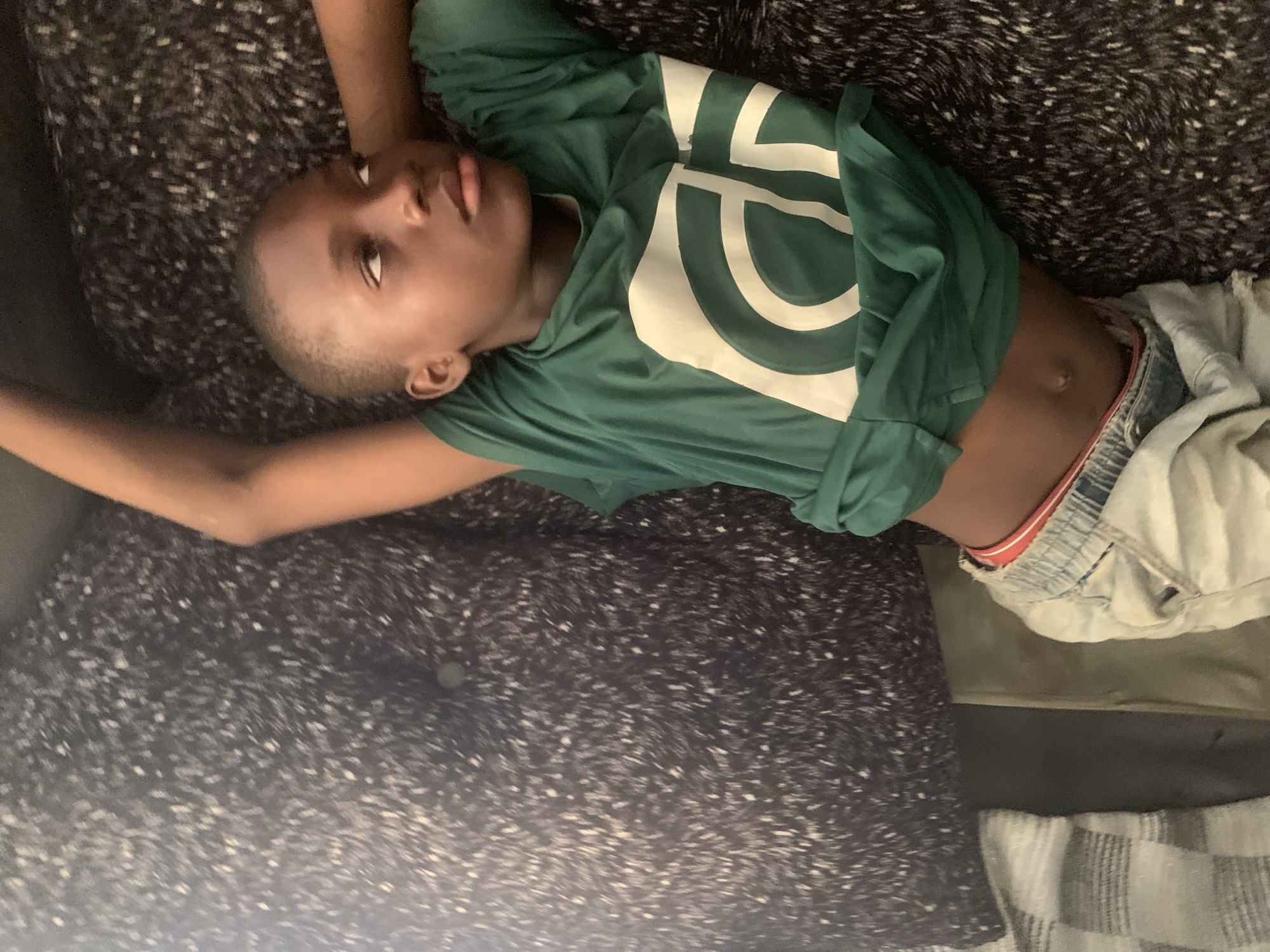 A young Black child is lying on their back on a textured surface, possibly a couch or bed. The child is wearing a green long-sleeved shirt with a large white graphic design on it. The shirt is pulled up slightly, revealing the child's abdomen and the waistband of their light-colored shorts, which have a red elastic band. The child's head is tilted back, and their eyes are open, looking upwards. Their left arm is extended above their head, while their right arm is bent at the elbow, resting on their body. The background is dark and textured, with a speckled pattern. The lighting appears to be artificial and dim, suggesting it might be evening or nighttime. There are no discernible weather cues or specific location details within the frame beyond the possibility of an indoor domestic setting. The child's expression is neutral. No visible text is present in the image.
