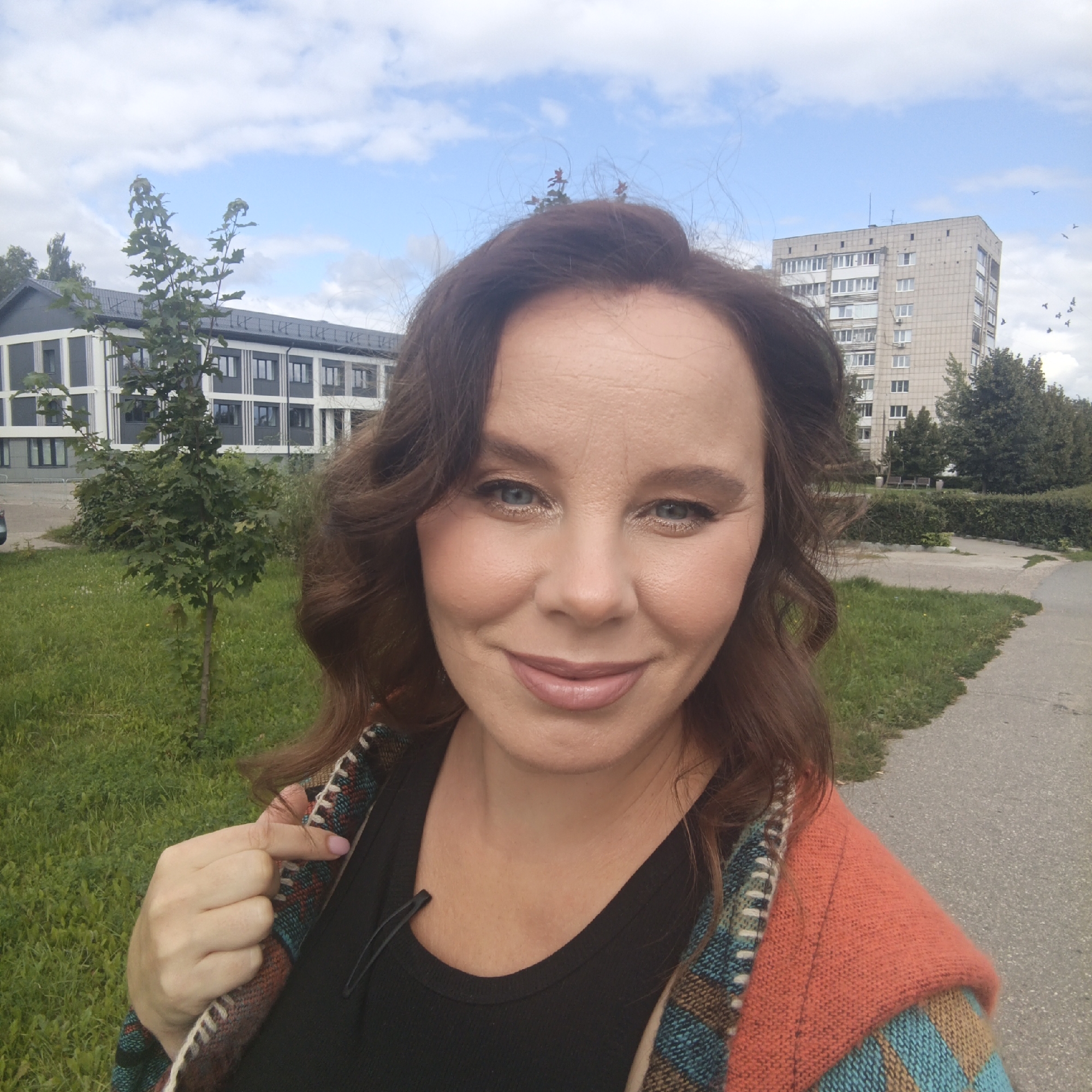 The image is a selfie taken outdoors, likely in a residential area of Chaikovsky, Russia, given the presence of typical Soviet-era apartment blocks and newer buildings. In the foreground, a woman with brown hair and makeup is captured from the chest up, smiling slightly. She is wearing a black tank top and a colorful knitted cardigan or shawl in shades of orange, blue, and brown. Her left hand is visible, touching the cardigan. In the background, two buildings are visible: a modern, multi-story building with a gray facade and dark windows on the left, and a taller, older, beige apartment building with many windows on the right. A small, young tree with green leaves stands in the grassy area between the woman and the modern building. A paved path runs along the right side of the frame, bordered by grass. The sky is blue with scattered white clouds, and a flock of birds is visible in the upper right portion of the image. The overall scene suggests an urban or suburban environment on a partly cloudy day.