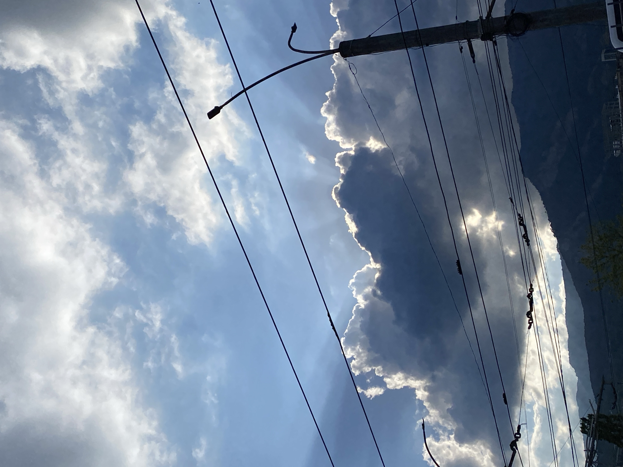 The image shows a view of the sky with clouds and power lines. In the background, there's a dark mountain ridge. A utility pole with a streetlight is visible, casting shadows on the clouds. The sky varies from light blue to dark gray due to the clouds. The scene appears to be set in a city with visible infrastructure, possibly in город Ялта, Ukraine, judging from the mountain landscape and urban elements. No people are visible.