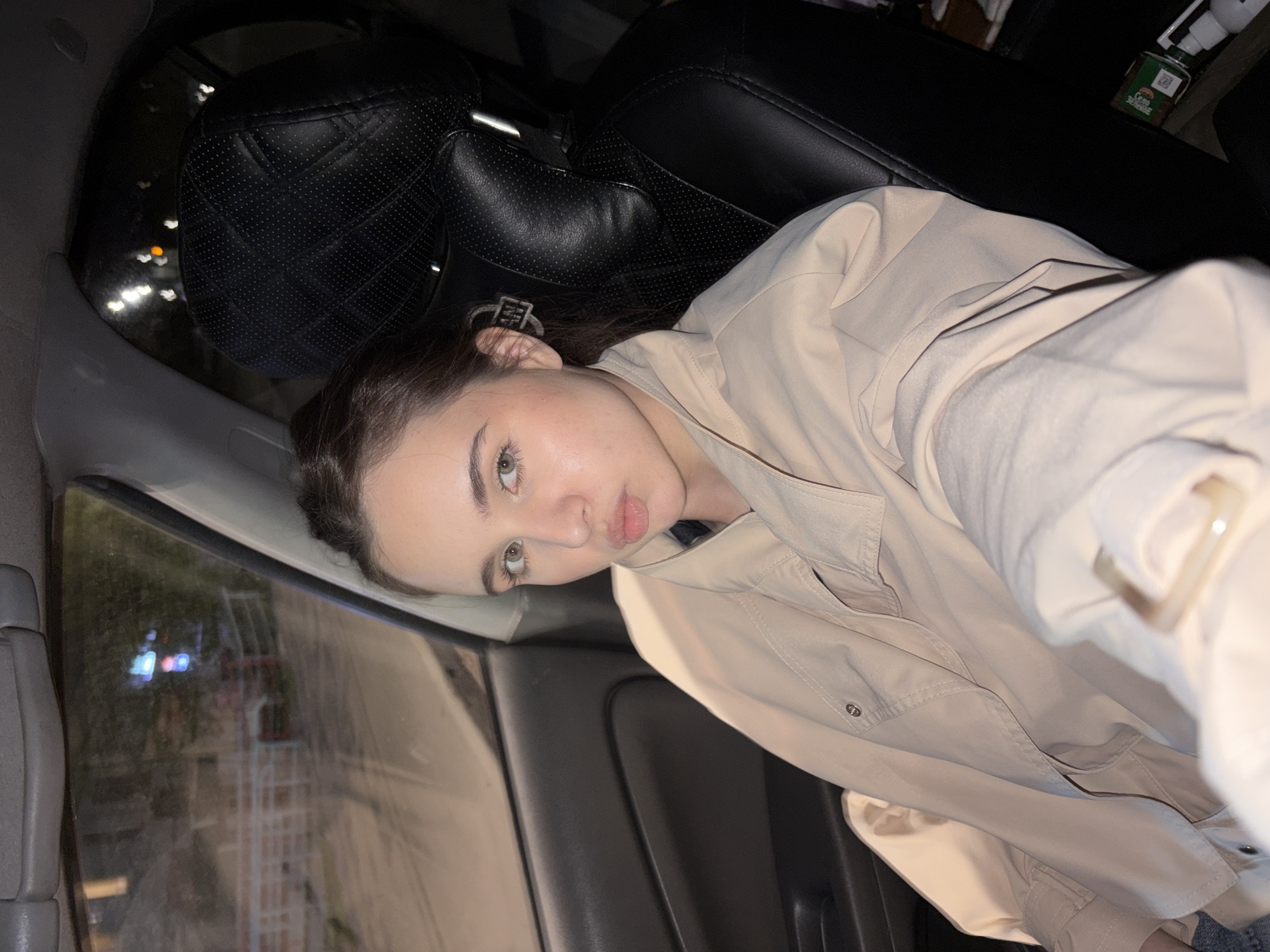 A young woman is in the passenger seat of a car. She has brown hair and green eyes, and is wearing a light tan jacket. The car's interior is mostly black, with gray trim on the door. Outside the window, streetlights and buildings are visible. The location is Izhevsk, Russia.