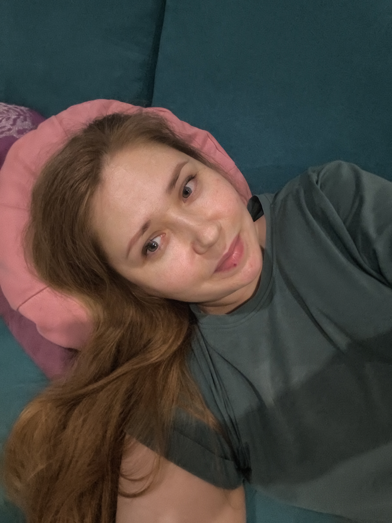 This is a selfie of a young woman with long, reddish-brown hair, lying on a teal-colored couch. She is resting her head on a pink pillow and a darker purple pillow. The woman is wearing a dark green t-shirt. She has light skin, light brown eyes, and a small mole on her chin. Her expression appears relaxed and content, with a slight smile. The lighting suggests it is daytime, possibly indoors, given the setting. No text is visible in the image. The location context provided is Kazan, Russia.