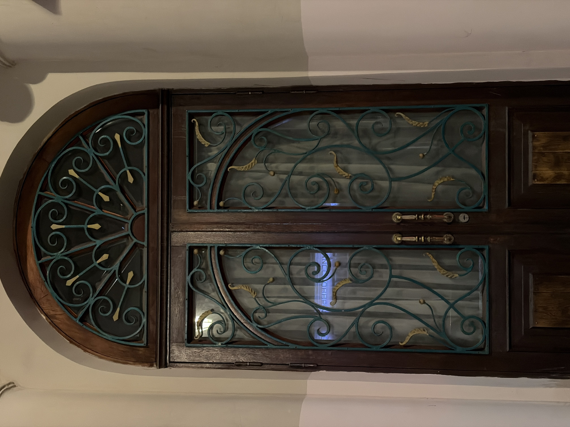 This is a full-frame, eye-level, indoor photograph of an ornate wooden double door with a semi-circular transom window above. The door is dark brown wood with visible grain. Both door panels have large glass panes framed by decorative teal-colored metalwork with gold accents. The metalwork features swirling filigree and leaf-like shapes. The transom window also has a similar metal grate design, resembling a fan or sunburst. The door has two brass door handles and a visible keyhole. The door is set into a neutral-colored wall, with a slight shadow cast on the right side of the frame. The scene is likely a building entrance in Saint Petersburg, Russia, given the architectural style and ornate details commonly found in the city.