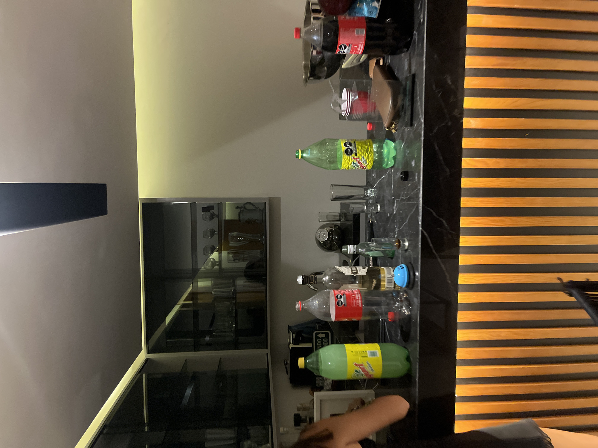 This indoor image, taken in San Bernardino Tlaxcalancingo, Mexico, captures a counter or bar area illuminated by artificial lighting, suggesting it is evening or night.
The central feature is a dark, possibly marble or granite counter laden with various bottles and drinkware. Several large beverage bottles are present: a red-labeled Coca-Cola bottle (visible text: "Coca-Cola," "Sin Azúcar") and two green-bottled sodas, one with a yellow label and drawn-on cartoon eyes (visible text: "CONT. NET."). Numerous smaller clear glass bottles, likely containing spirits, are also arranged on the counter; text "TRES" is visible on one bottle, and "SMIRN..." (possibly Smirnoff) on another. Shot glasses, a red plastic cup, and a metal bowl are interspersed among the bottles. A dark brown wallet or clutch rests on the counter.
To the left, a glass-fronted cabinet or display unit holds an assortment of glasses and other items. A dark, rectangular object, possibly a wall-mounted fixture, is visible higher on the left wall, which is light grey. The wall on the right side of the frame is a yellowish-beige.
On the far right, a decorative wall panel features horizontal wooden slats against a dark background. In the lower right corner, the arm of a person, wearing a dark sleeve, is visible reaching towards the counter, suggesting interaction with the drinks. The scene depicts a setup for beverages, likely for a social gathering or home entertainment.