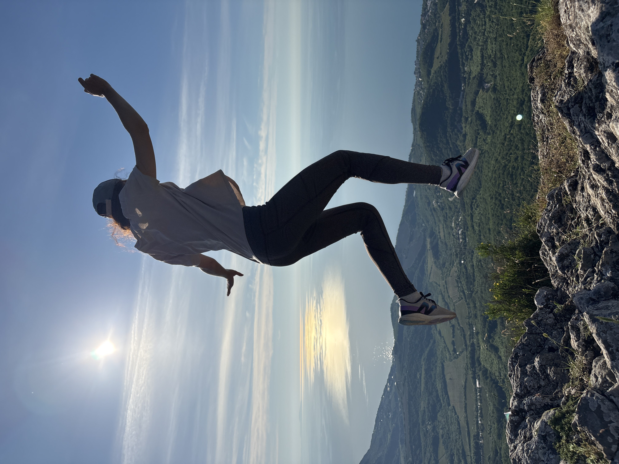 An individual is captured mid-jump against a luminous sky and coastal panorama in the city of Unknown, Ukraine. The subject, viewed from the back, is silhouetted and appears to be in an upward motion, with arms extended and bent, hands open, and legs bent. They are dressed in a light-colored t-shirt, dark textured leggings, and athletic shoes with white, grey, purple, and blue accents. A dark baseball cap is worn backward. The sun is prominently visible in the upper left, creating a strong backlight and a brilliant lens flare. Below the subject, a rocky foreground with sparse dry grass and small plants gives way to a vast midground of undulating green mountains and valleys, dotted with small towns and settlements along a coastline. The background features a wide expanse of calm, blue sea or ocean, reflecting the sun in a shimmering, golden path that extends towards the horizon. The sky above is clear blue with several thin, wispy white clouds.
