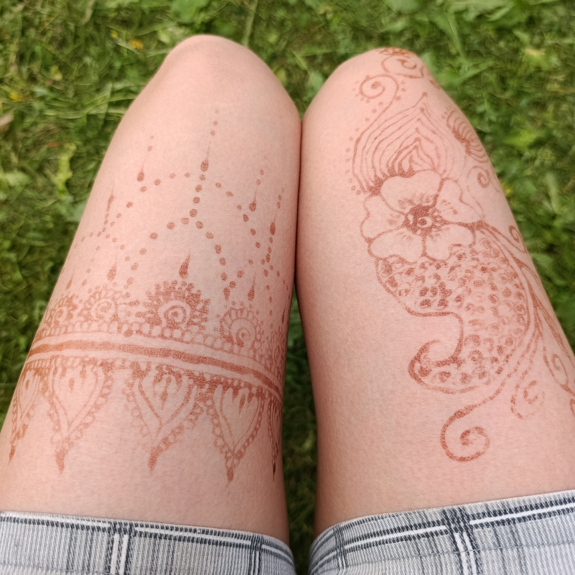 The image shows a close-up view of two legs with henna tattoos. The legs are positioned vertically in the frame, with the thighs visible from the knees upwards. The skin is fair and smooth, and the henna designs are a reddish-brown color. On the left thigh, a decorative band with intricate patterns is visible, resembling lace or tribal markings. There are also scattered dots and teardrop shapes above this band. The right thigh features a larger, more elaborate design, including a stylized flower with petals and a detailed, textured center. Swirling lines and smaller floral elements complete the design on the right leg. The subjects are wearing light-colored shorts with a checkered pattern. The background consists of green grass, out of focus, suggesting an outdoor setting. No specific location in Dubovaya, Russia, can be determined from the image.
