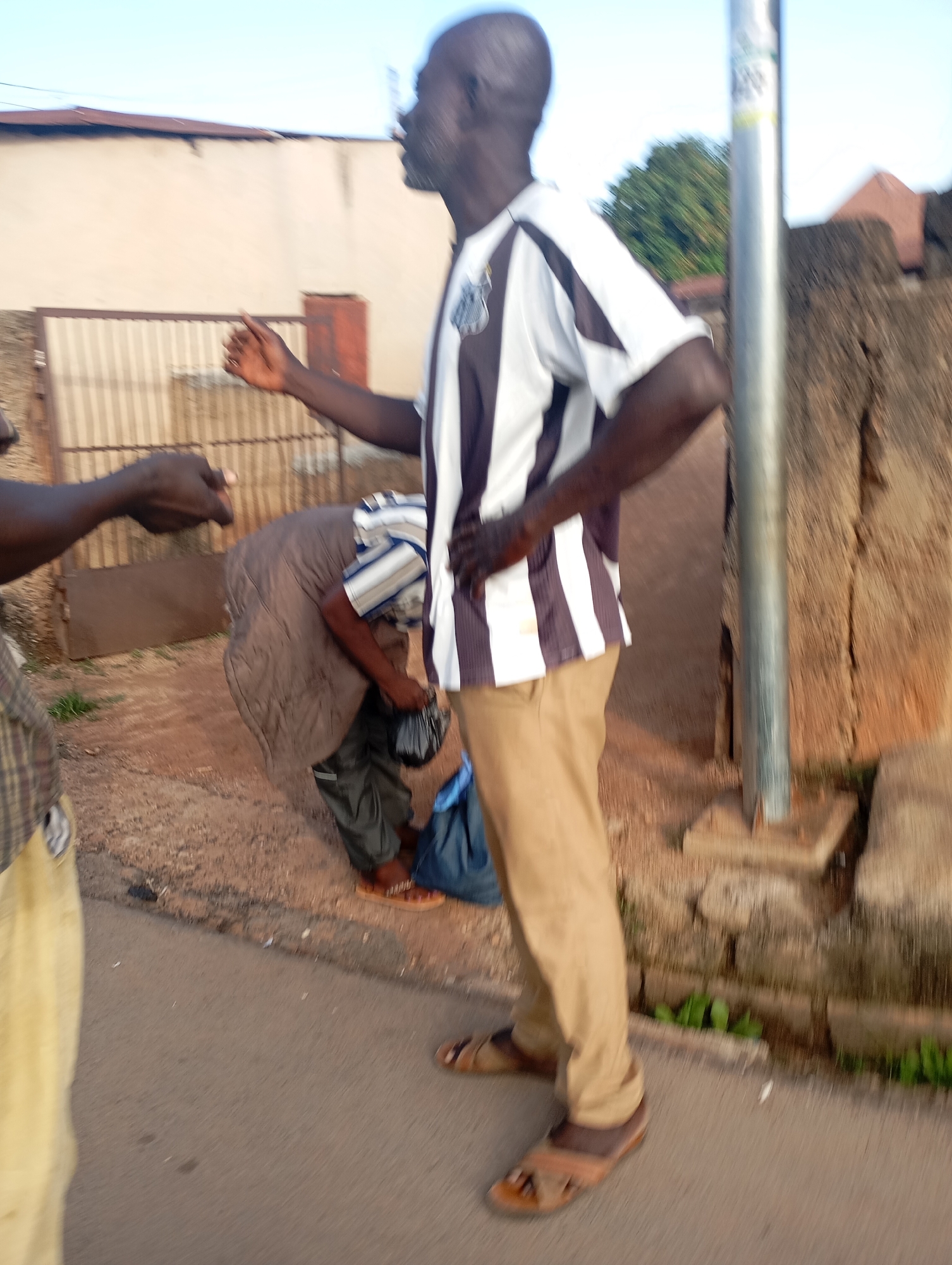 The image depicts an outdoor scene, likely in the city of Shendam, Nigeria. A man wearing a black and white striped shirt and tan pants stands on the right side of the frame, facing left. His right hand is resting on his hip, and his left arm is extended slightly forward. To his left, another person is bent over, seemingly interacting with or picking up items from a blue plastic bag on the ground. This person is wearing a striped shirt and dark pants. In the background, a beige building with a corrugated metal fence is visible. A metal pole stands vertically in the foreground to the right of the central figure. The ground is a mixture of dirt and asphalt.