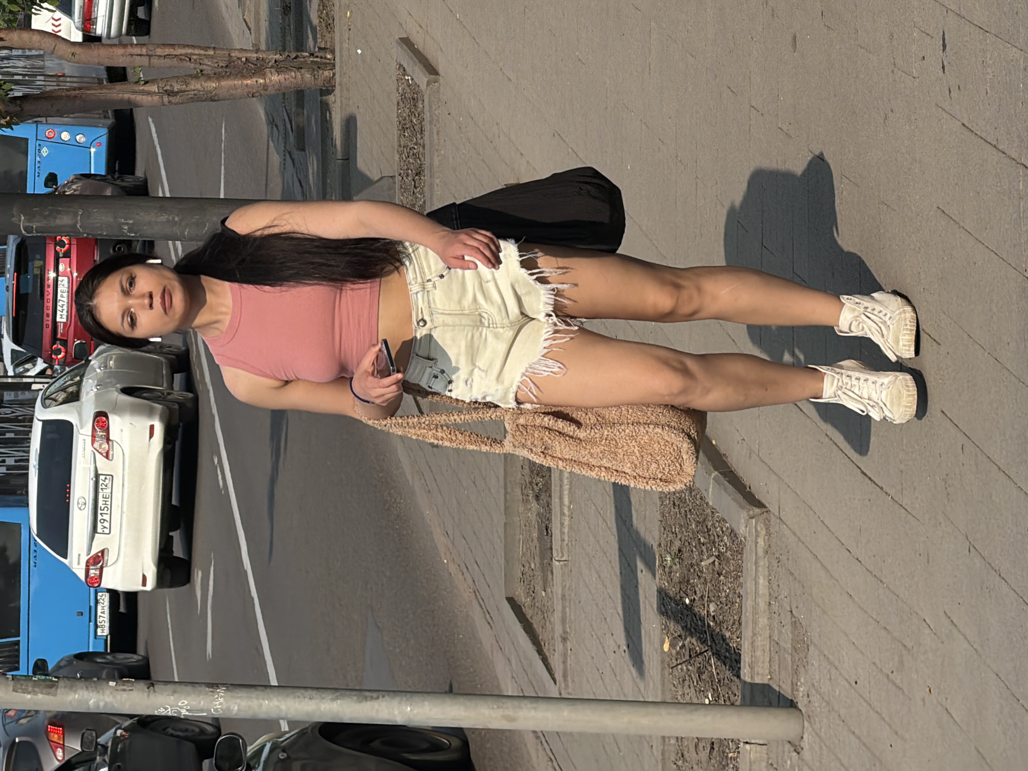 A young woman with long dark hair and white earbuds walks on a sidewalk in Krasnoyarsk, Russia. She is wearing a pink crop top, distressed denim shorts, and white high-top sneakers. She carries a beige furry tote bag on her left shoulder and a black bag on her right. The sidewalk is paved with concrete slabs, and there is a raised planter bed with dirt and some debris to her right. Behind her, a street with several vehicles is visible, including a white Toyota sedan with a license plate ending in "124", a blue bus, and a red SUV. The scene is illuminated by bright sunlight, casting shadows on the ground. The background appears to be an urban environment.