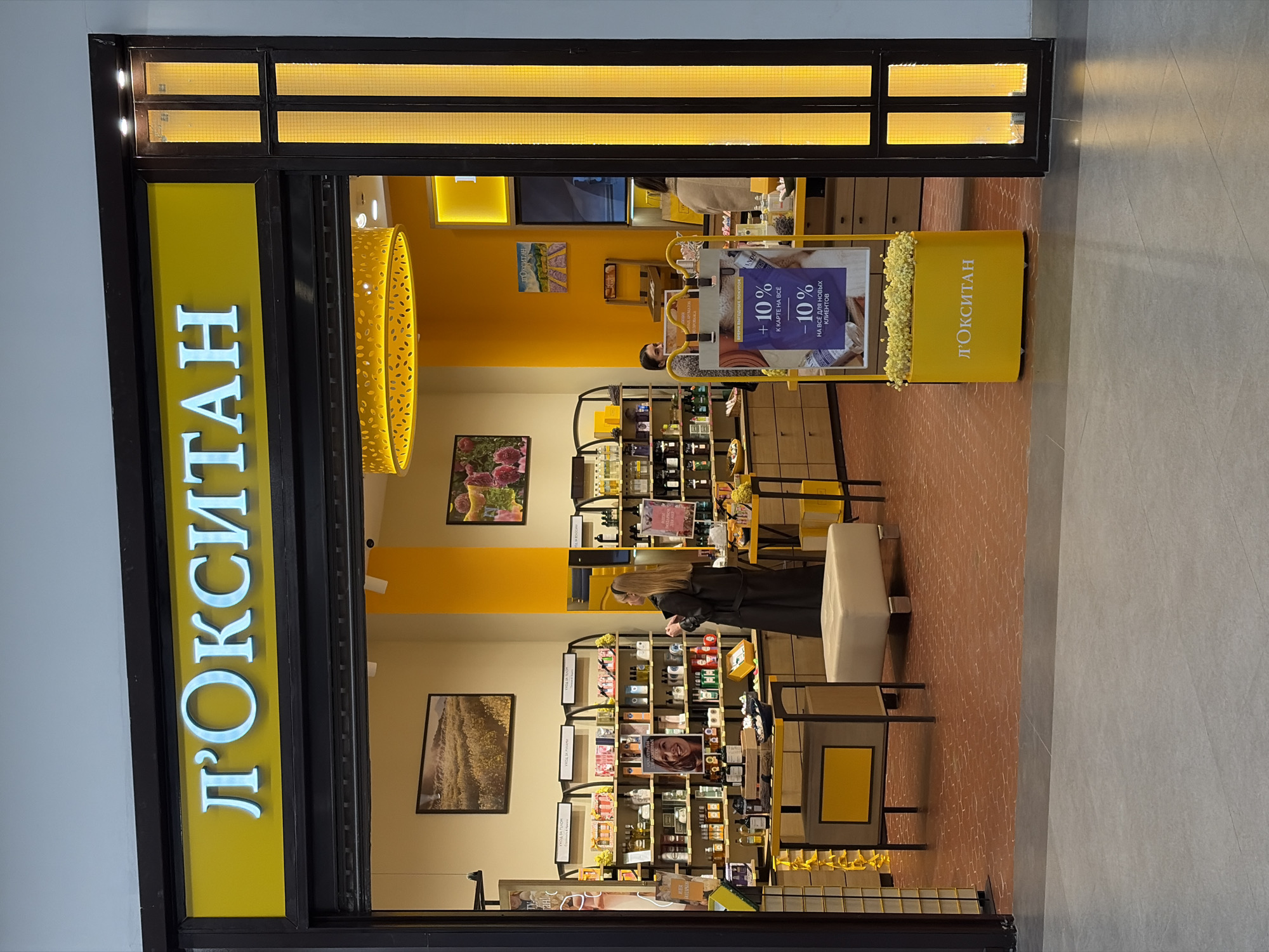 The photograph captures the entrance of a L'Occitane store, likely located in the city of Izhevsk, Russia. The sign above the entrance prominently displays the store's name in Cyrillic letters, colored in luminous white against a yellow background. The storefront is framed by a dark structure with a rectangular grid of lights above the entrance.
Inside the store, the interior is painted in a light yellow hue, creating a bright and inviting atmosphere. Shelves are filled with a variety of L'Occitane products, meticulously arranged for display. A woman is seen examining the products on one of the shelves, suggesting customer interaction.
In the store, promotional signage with the text "+10% to the card for everything" and "-10% for new customers" can be noticed. A pendant light with a geometric design hangs from the ceiling, adding a touch of modern elegance to the space.
The flooring inside the store is patterned with a dark yellow-orange stone pattern. The shop is situated on a stone-tiled floor. The photograph is taken from a slightly elevated angle, providing a clear view of the store's entrance and interior.