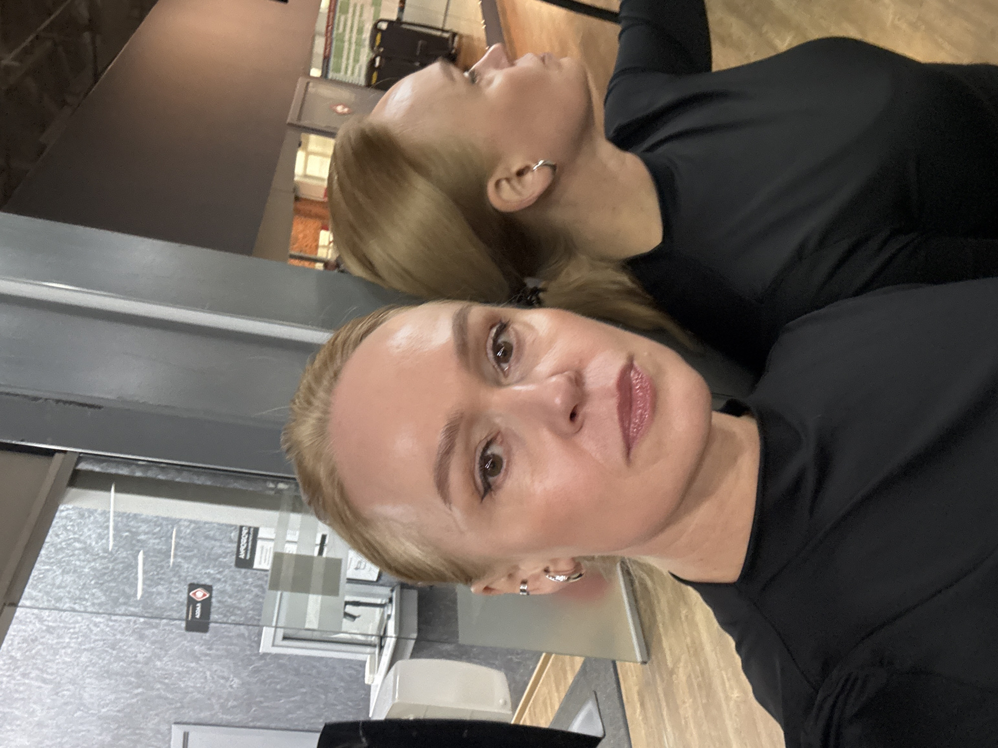 A woman in a black long-sleeved shirt is taking a selfie in what appears to be a medical office or beauty salon. Another woman is visible slightly out of focus behind her, also wearing a black top. The background includes a waiting area with chairs, a reception desk, and various posters and signage. The location is possibly in Chaikovsky, Russia.