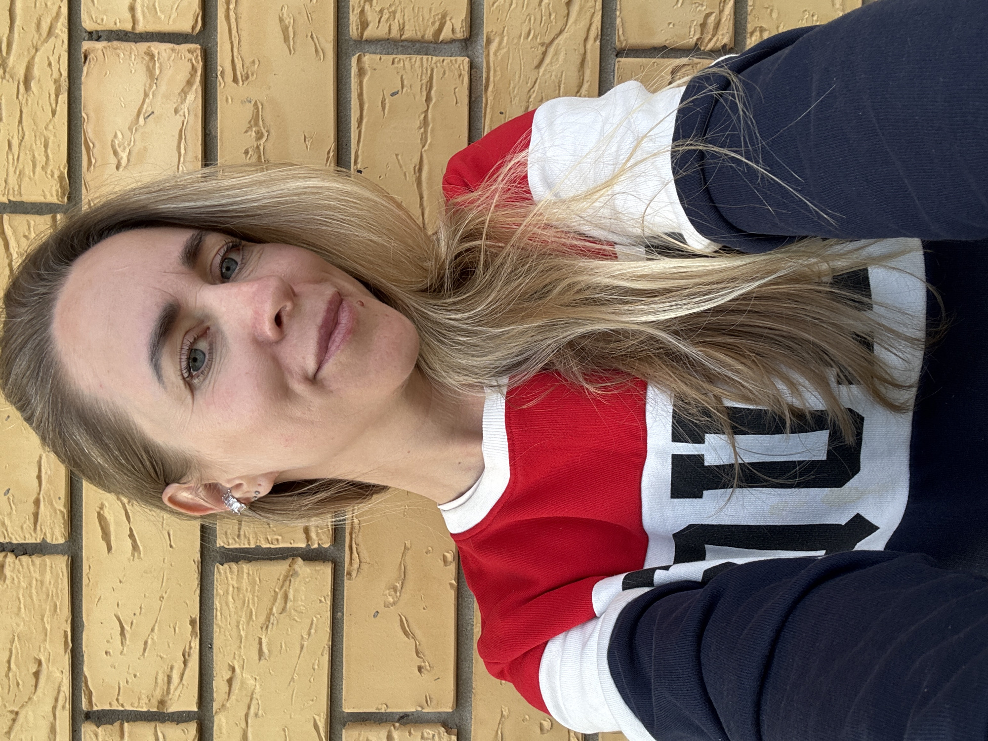 A woman with long, blonde hair and blue-green eyes is captured in a close-up selfie, looking directly at the camera with a slight, calm smile. She appears to be an adult, likely in her 30s or 40s. Her fair skin shows minimal makeup. On her visible left ear, she wears a silver-toned earring or ear cuff with small, possibly sparkling, details.
She is wearing a long-sleeved top that features a color-blocked design in red, white, and dark blue (or navy). The left side of her torso and shoulder is red, the central part with the visible text is white, and her right sleeve and lower torso are dark blue. Large, bold black block letters are prominently displayed on the white section of her top. The letter "P" is clearly visible, followed by what appears to be part of another character, possibly "1" or "O", forming a logo or design.
The background is a textured brick wall, featuring yellowish-brown or tan bricks with dark gray mortar lines. The rough surface of the bricks indicates an outdoor setting, consistent with the bright, natural daylight illuminating the scene. The overall impression is a casual portrait taken in Chaikovsky, Russia.