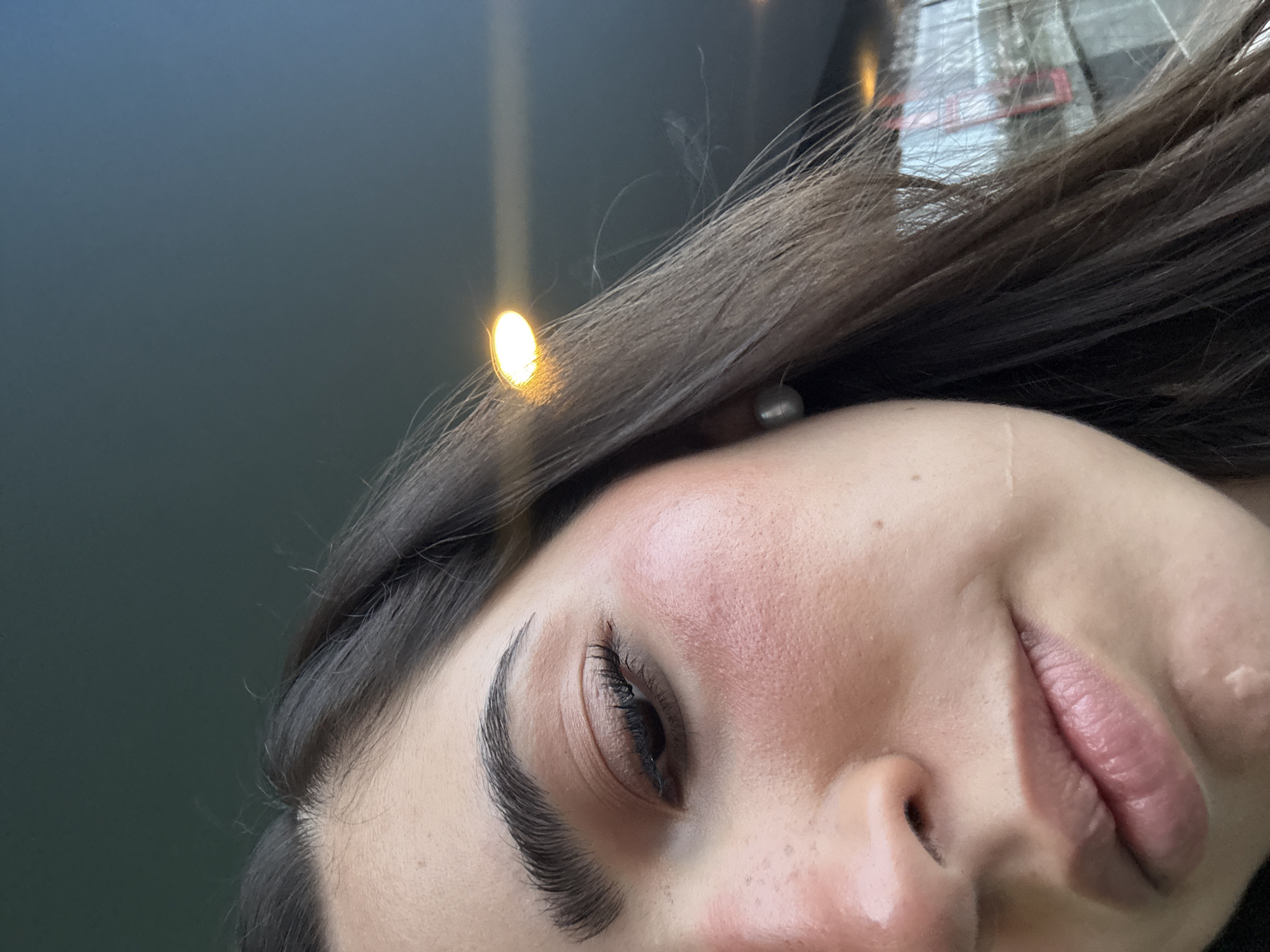 The media file is a close-up, low-angle shot of a person's face, primarily focusing on the right side. The subject, likely female, has dark brown hair that falls across the forehead. Visible features include the right eye with long, dark eyelashes and a sculpted eyebrow, a cheek with a reddish flush, the nose, and a portion of the lips, which show a subtle upward curve at the corner. A small, light-colored stud earring is visible near the hairline on the right side.
The image is taken from a perspective looking up at the person. The background is a dark, muted blue-grey, suggesting an indoor setting or an evening/night outdoor scene. Several distinct, bright, circular yellow-orange light reflections are present, particularly in the upper left and center-right. In the upper right corner, there are blurred reflections that resemble urban structures or buildings, including a noticeable red element, possibly indicating a window, sign, or part of a building.
The subject's expression appears neutral or slightly pleasant. There is no discernible activity other than being photographed in this close-up. The lighting, characterized by bright reflections, points towards artificial light sources. No text is visible in the image. The location context is San Bernardino Tlaxcalancingo, Mexico.