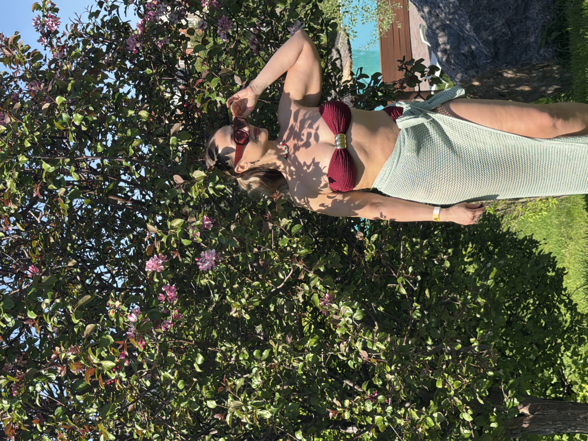 The image displays a woman reclining outdoors under bright sunlight, within a lush, green setting. She is wearing a dark red, bow-shaped bikini top with a prominent gold-colored metal clasp at the center, paired with a light mint-green, knitted sarong or wrap skirt tied around her waist. Her red-framed sunglasses are pushed up onto her forehead, and her right hand is casually placed near them. She also wears a necklace with red and turquoise beads and a gold-colored bracelet on her left wrist.
The woman is positioned among dense foliage of a flowering tree or large bush, which features both green and reddish-brown leaves and small pink blossoms. The tree occupies a significant portion of the left side of the frame. The strong, direct sunlight casts distinct shadows across her body and the surrounding environment, indicating a clear, sunny day, likely during midday or early afternoon. In the background, to the right, glimpses of a bright blue swimming pool, green grass, and a large natural rock or stone are visible. The scene suggests a relaxed atmosphere, possibly a private garden or poolside area located in Krasnoyarsk, Russia. The woman appears calm and at ease, enjoying the sun. No text is visible in the image.