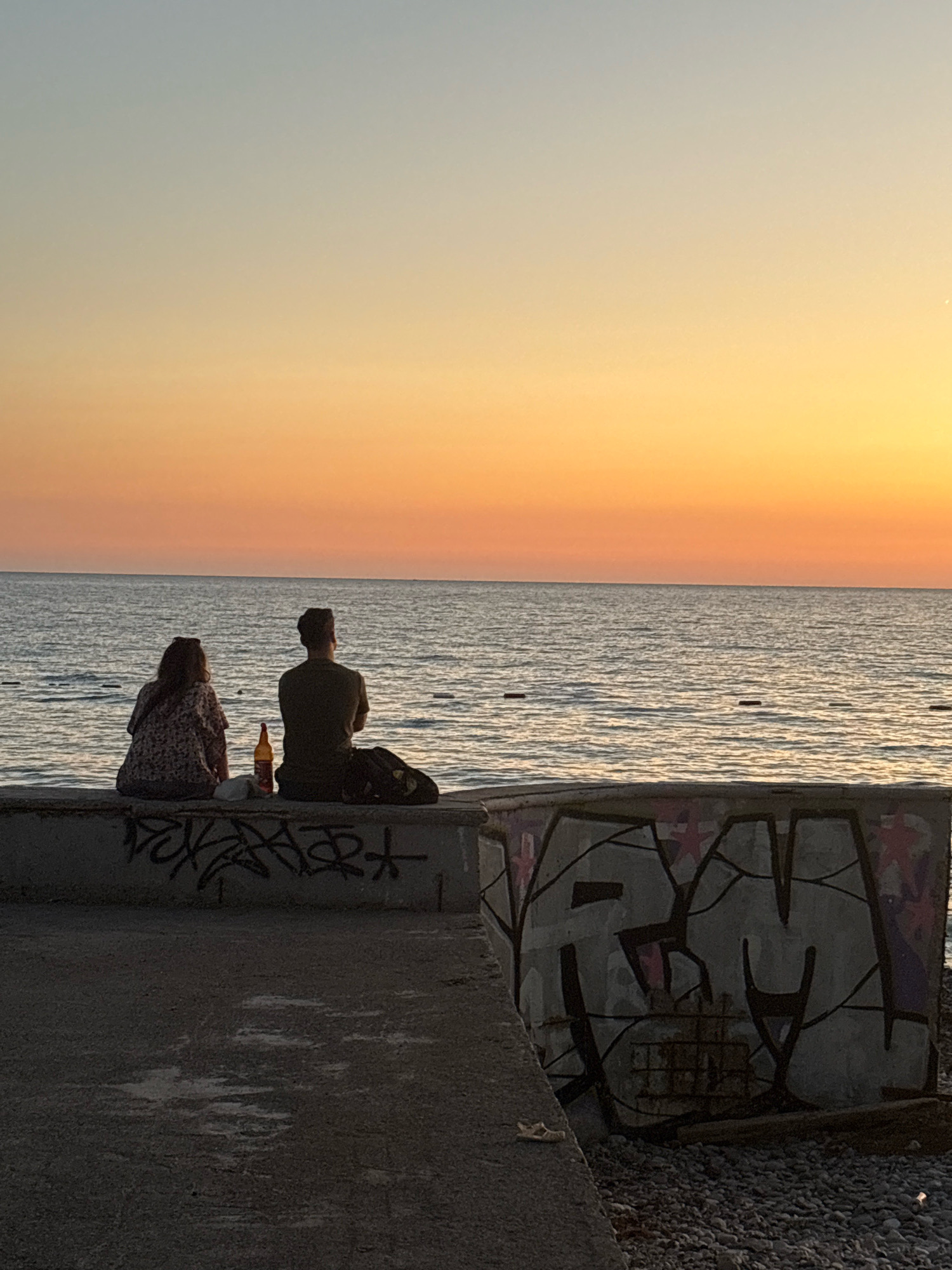 The image depicts a serene sunset scene at the Bar coast in Montenegro. Two individuals, a male and a female, are seated on a concrete barrier overlooking the sea. The female, on the left, is wearing a patterned blouse and sunglasses. The male, on the right, is in a dark t-shirt and has a backpack next to him. A bottle of beer stands between them on the barrier. The concrete barrier itself is adorned with graffiti. The sea is calm, reflecting the warm hues of the sunset, which transitions from a soft orange near the horizon to a pale blue in the upper sky. The foreground shows a textured concrete walkway leading to the barrier.