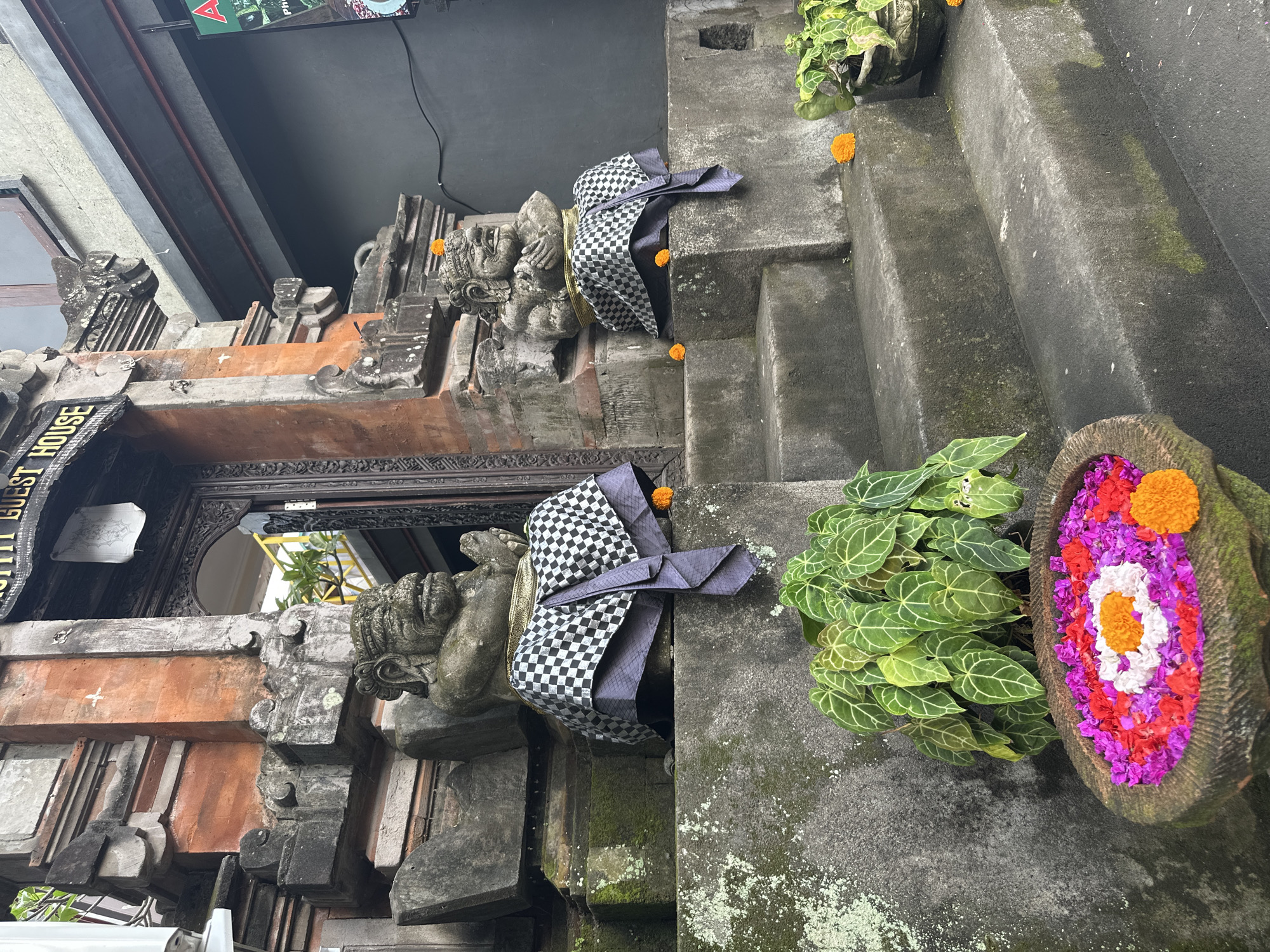 The image shows an outdoor scene in the Ubud District, Indonesia, likely in front of a guesthouse. Two stone statues, resembling gargoyles or mythological figures, sit on either side of a set of stone steps. These statues are adorned with black and white checkered cloths and are decorated with small orange flowers. In the foreground, a large stone bowl filled with vibrant, colorful flower petals, including pink, purple, red, and white, with a prominent orange and white flower in the center, sits next to a lush green plant. The steps themselves are made of stone and show signs of age and moss growth. Behind the steps and statues, a traditional Balinese architectural entrance is visible, featuring intricate wooden carvings and brickwork. The overall impression is one of a culturally rich and spiritual location, possibly a place of worship or a traditional Balinese dwelling.