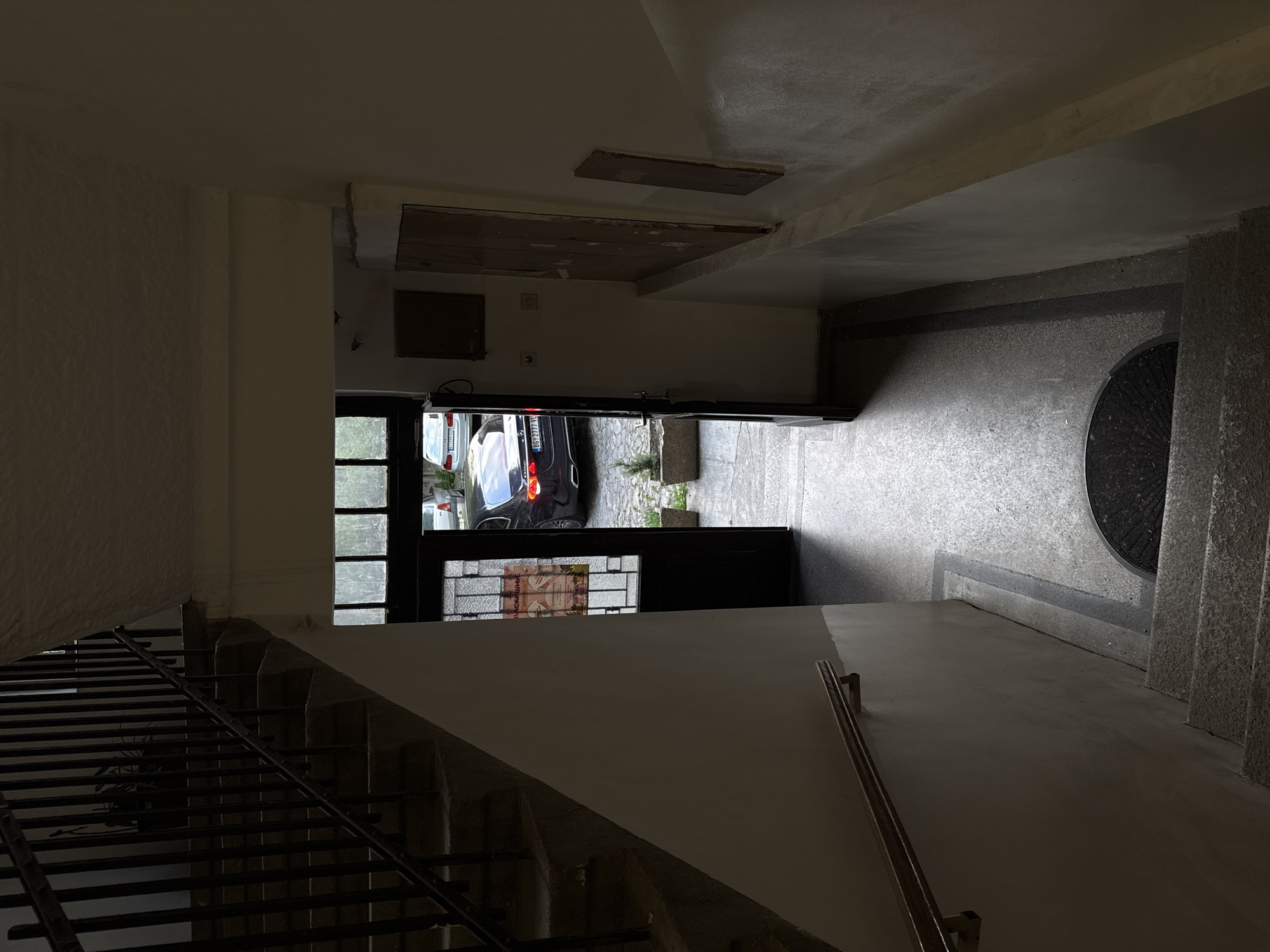 The image shows a view from inside a stairwell looking out through an open doorway. The stairwell has concrete steps with a metal railing. The floor outside is paved with a doormat visible. A black car is parked partially visible through the doorway.