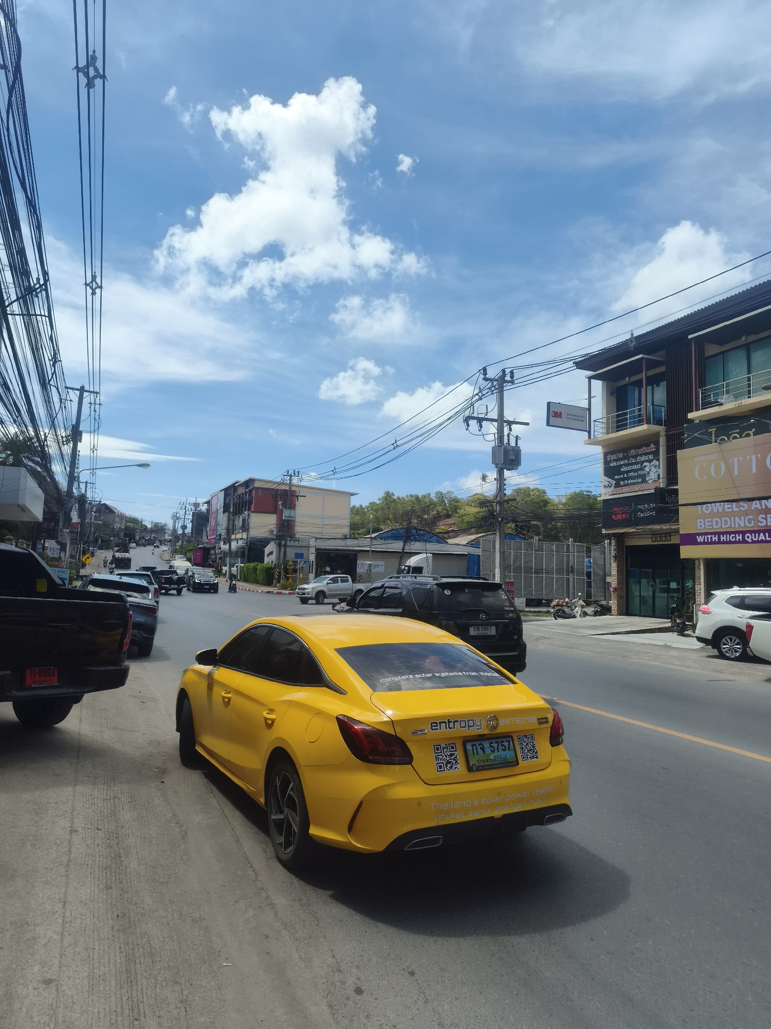The image shows a street scene in the Ko Samui District of Thailand under a partly cloudy blue sky. A bright yellow car, identified as an MG, is parked on the side of the road, facing away from the camera. The car has several decals on its rear, including one that reads "entropy" and a license plate with Thai characters and numbers "ตจ 5757". Another decal on the car states "Thailand's solar power leader phuket, samui, phangan, hua hin".
Several other vehicles are visible on the road, including a black pickup truck in the foreground to the left and a black SUV to the left of the yellow car. A white SUV is parked further down the street on the right. Commercial buildings line the right side of the street, with signs in Thai and English. One sign indicates "COTTON TOWELS AND BEDDING". Utility poles with a dense network of electrical wires dominate the left side of the frame. The road is paved and has a yellow line marking the lane division. The general environment appears to be a commercial area with a mix of businesses and vehicles.
