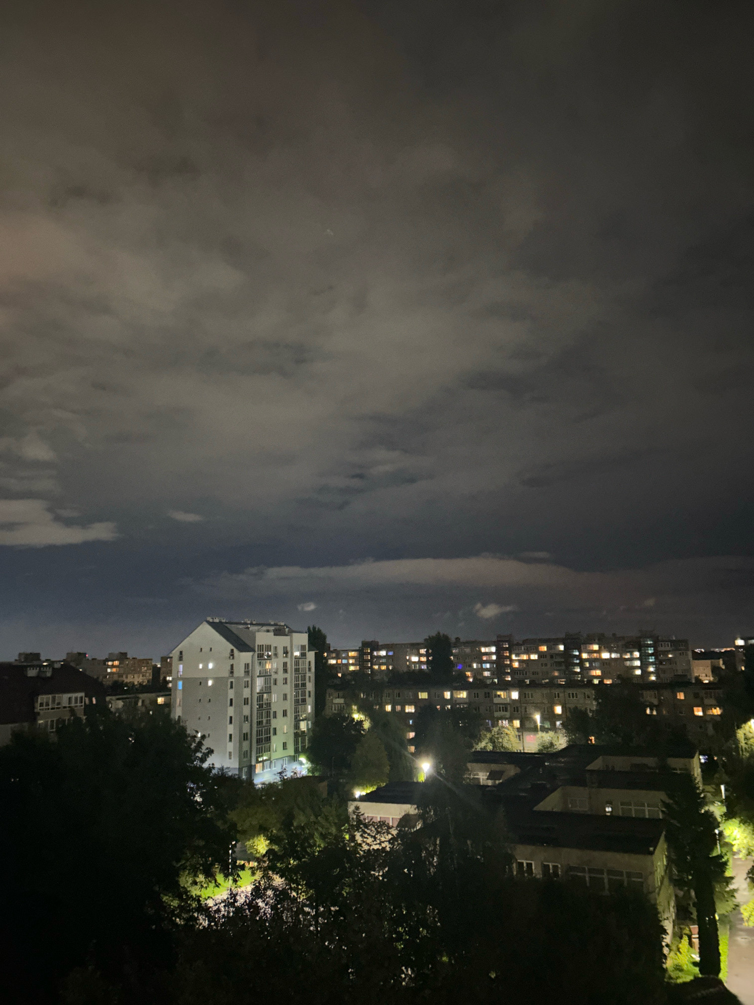 An outdoor, elevated nighttime shot captures a cityscape under a cloudy sky. A tall, modern white building with many lit windows stands prominently in the middle ground, its facade illuminated against the dark sky. Behind and to the sides of this building are other structures, including lower apartment blocks with numerous glowing windows, suggesting residential activity. Trees with dark foliage are visible in the foreground and between the buildings, some of them illuminated by artificial lights, creating patches of light and shadow. The sky is filled with gray, textured clouds, some darker and more ominous than others, hinting at a potentially stormy or overcast condition. The overall scene depicts an urban environment at night, characterized by the interplay of artificial lights from buildings and the natural darkness of the night sky. The location is the city of Kaliningrad, Russia.