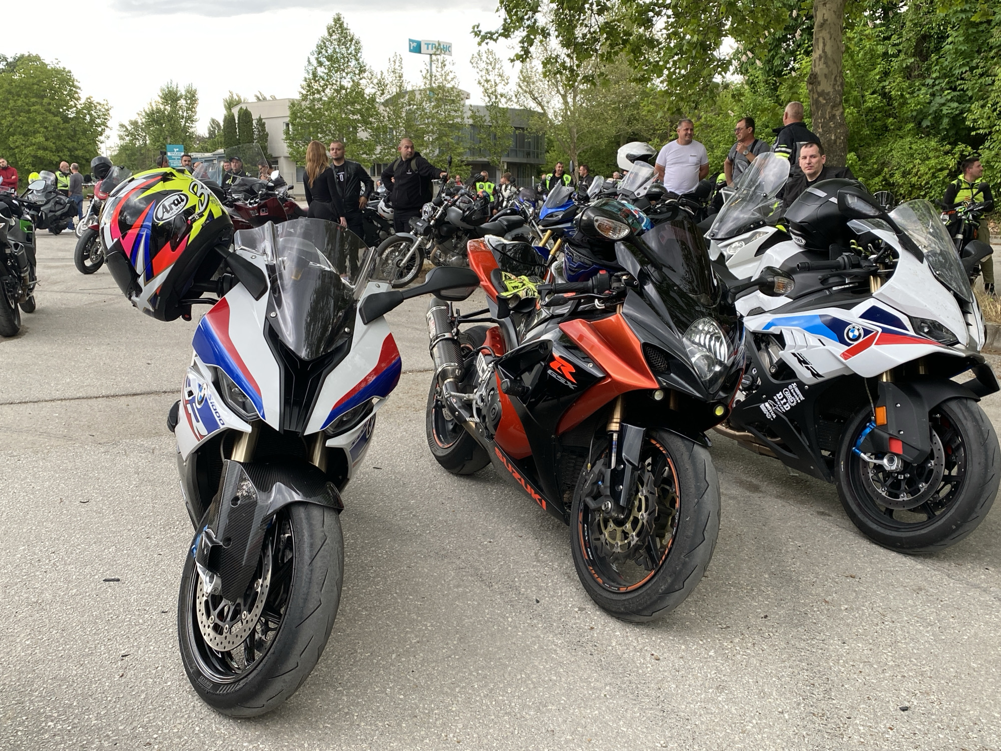 The image shows a gathering of motorcycles and people on a paved surface, likely a parking lot, in Pleven, Bulgaria. In the foreground are three motorcycles: a white and blue BMW S1000RR with a helmet resting on its handlebar, a black and orange Suzuki GSX-R, and another white and blue BMW S1000RR. Behind the motorcycles, a group of people are standing and talking. In the background, a building with a sign bearing the name "TRANC" can be seen amongst the trees. The sky is overcast.