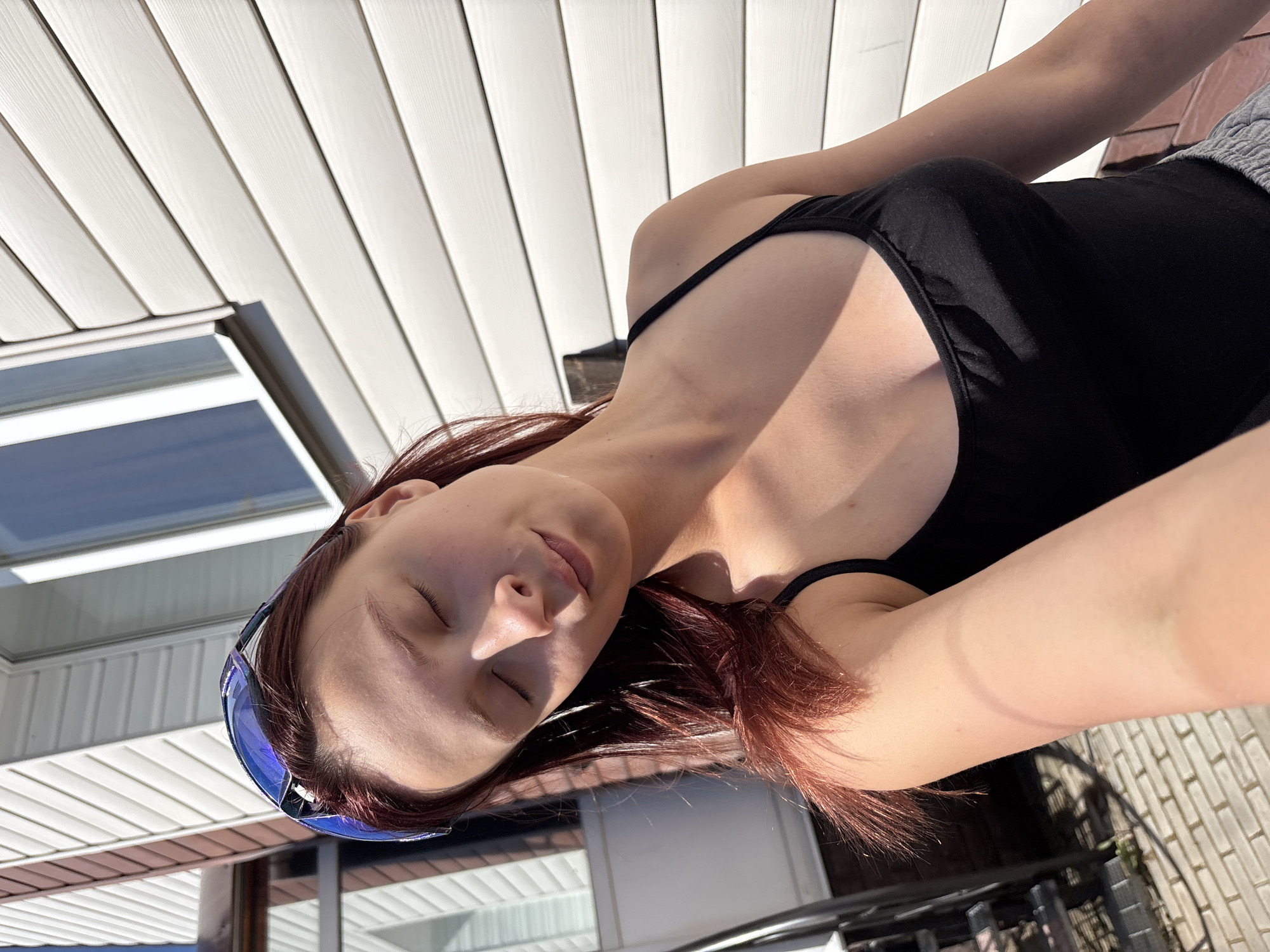 The image shows a young woman with reddish-brown hair wearing a black tank top and grey sweatpants. She is outdoors, leaning against a white vinyl-sided building. Blue-tinted sunglasses are perched on top of her head. Her eyes are closed, and she appears to be enjoying the sunlight. The background includes a brick-paved area and what looks like stairs. The environment suggests a residential setting, possibly a backyard or patio. The location is specified as Прикамский, Russia.