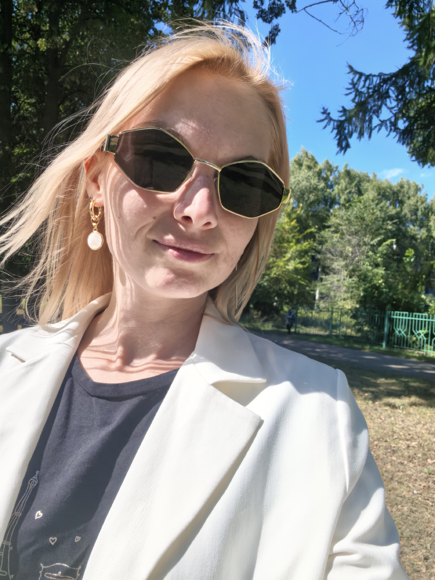 A woman with blonde hair and sunglasses is taking a selfie outdoors. She is wearing a white blazer over a black t-shirt. She has a white circular earring with gold detailing. The background shows trees, a blue sky, and a green fence. The scene appears to be in a park or wooded area. The lighting suggests it is daytime with bright sunlight. There is no indication that this location is in Sterlitamak, Russia.