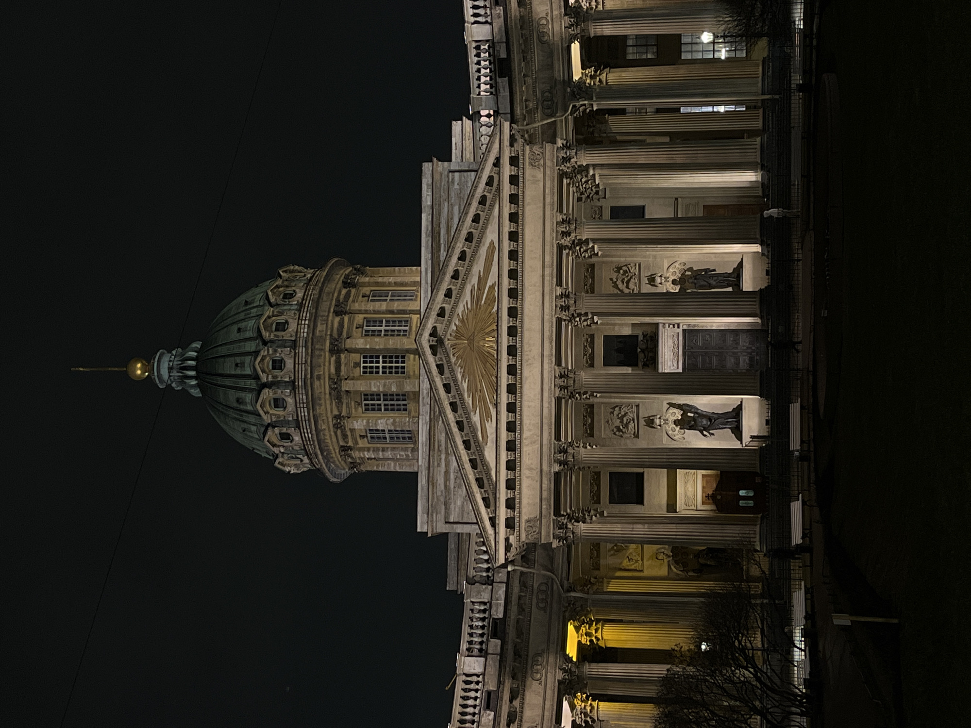 A nighttime shot of the Kazan Cathedral in Saint Petersburg, Russia, viewed at an extreme angle. The dome and facade are illuminated, showcasing architectural details such as columns and statues. Dark sky surrounds the structure. A lone wire extends from the dome. Dim lighting is seen through windows and along the building's lower edges. The foreground is obscured by darkness, with silhouettes of trees and benches.