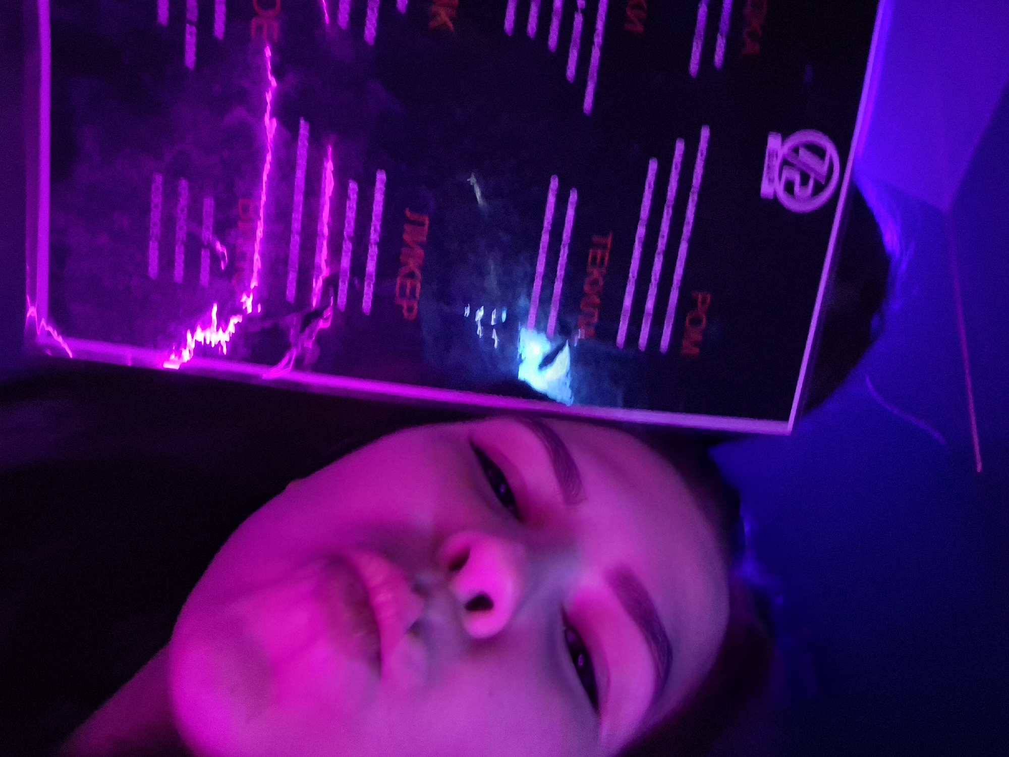 The media file captures a close-up, low-angle selfie or portrait of a person in a dark, neon-lit indoor setting, likely a bar or club, in Tobolsk, Russia. The dominant light source is magenta and purple neon, which intensely illuminates the person's face and a transparent, glowing menu board positioned above their head.
The subject is a young person, whose face is tilted, looking towards the camera with a neutral to slightly contemplative expression. Their features are highlighted by the vibrant pink and purple glow, casting some shadows.
Above the person, a transparent acrylic or glass board serves as a menu, with text and lines etched or printed in a way that allows them to glow in the same magenta hue. Visible Cyrillic text includes items commonly found on a drinks menu: "ЛИКЕР" (LIQUOR), "ТЕКИЛА" (TEQUILA), and "РОМ" (RUM). Partially visible words suggest "ВОДКА" (VODKA) and "ВИСКИ" (WHISKEY) or "КОКТЕЙЛИ" (COCKTAILS). A stylized circular logo, possibly "JR," is visible in the upper right corner of the menu board. Reflections and glare are prominent on the board's surface due to the intense lighting.
The overall atmosphere is dark and atmospheric, characteristic of a nightlife venue. The time of day is clearly nighttime or an indoor environment designed to appear so.