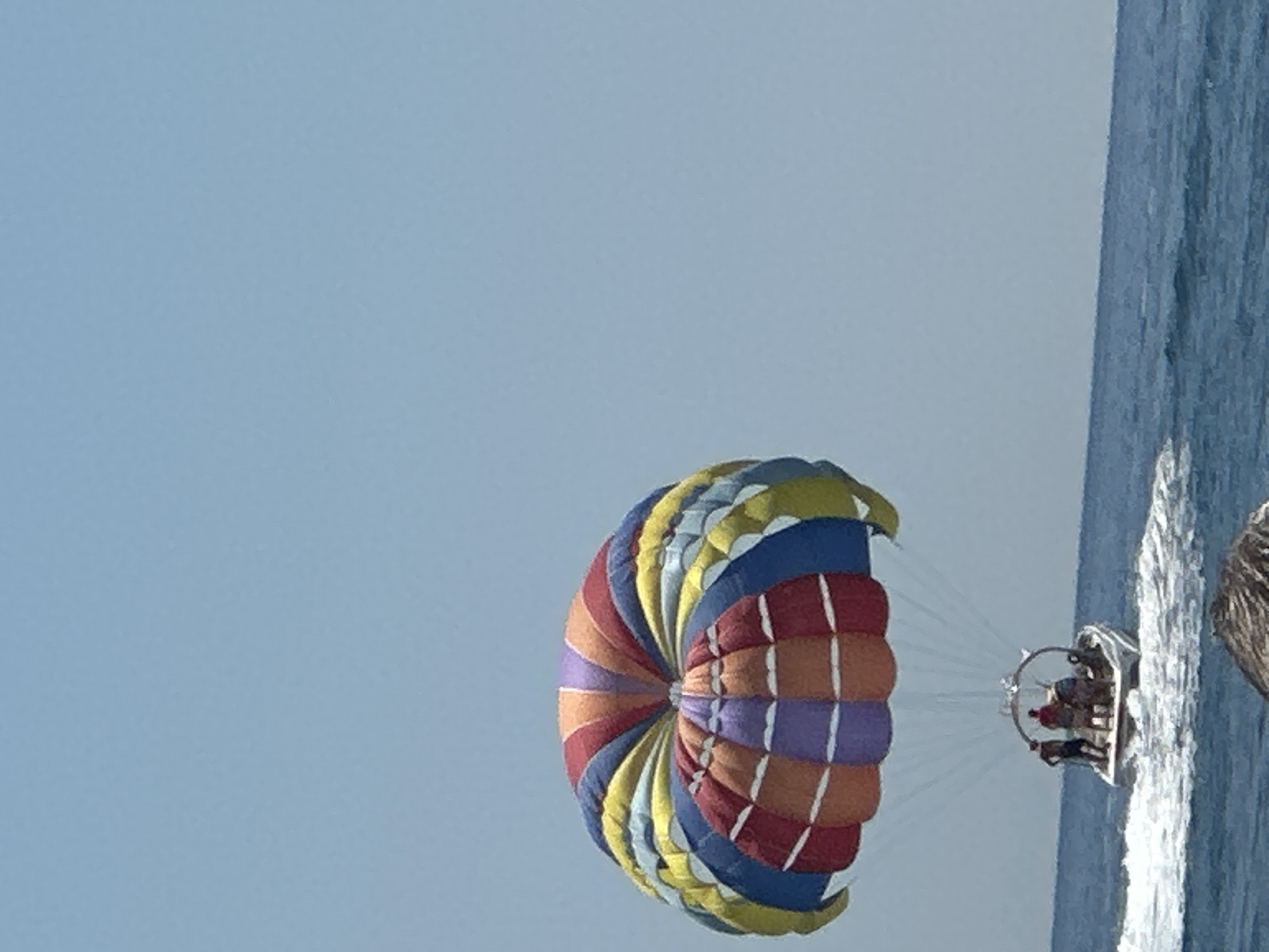 A colorful parasail with red, orange, purple, blue, yellow, and white panels is in the sky above a white boat on a blue ocean. Three people are visible in the boat, and the boat is creating white wakes in the water. The sky is clear and light blue. The image appears to be taken from another boat, with a blurry object in the foreground. The scene is consistent with parasailing activities that might occur in coastal areas like Boreti, Montenegro.