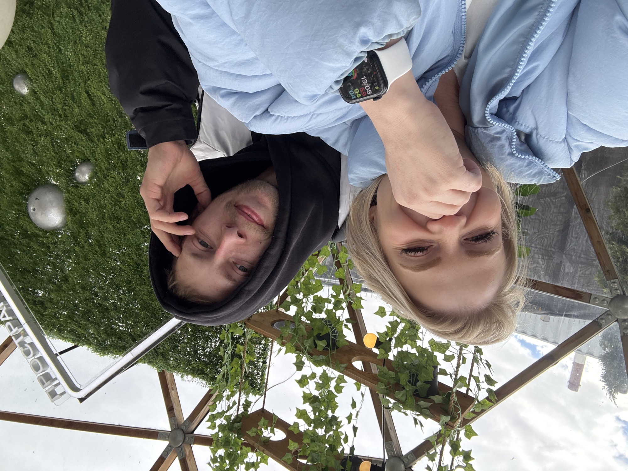 This media file captures a low-angle selfie of two individuals, a man and a woman, set within what appears to be a clear-roofed structure, possibly a greenhouse, gazebo, or an enclosed seating area. The image is oriented vertically, with the subjects appearing upside down relative to a typical upright view.
The subjects are positioned close to the camera, looking upwards directly at it. The man, on the left, has short facial hair and wears a black hoodie over a light-colored top. The woman, on the right, has blonde hair and is wearing a light blue puffer jacket. Her left hand is raised near her face, and on her left wrist, she wears a white-strapped smartwatch. The watch face is visible, displaying the time as 13:08 and the date as 04/04, along with some activity data. Both individuals exhibit neutral to slightly smiling expressions.
The setting appears to be a blend of natural and artificial elements. Behind the man, an area covered in artificial green turf is visible, featuring several metallic, spherical decorative objects. Above the subjects, visible through the clear roof panels, is a light grey, partly cloudy sky. A wooden framework adorned with artificial leafy vines and small yellow lights supports the transparent roof. In the distance, through the transparent panels, a brick chimney or tower can be glimpsed, suggesting an urban or industrial backdrop.
Visible text includes "ПРОСТРАН" on a white, rectangular sign, which is part of the Russian word for "space" or "area" (Пространство). The time of day indicated by the smartwatch is early afternoon. The overall impression is a casual moment captured in a unique, decorative outdoor or semi-outdoor space in Saint Petersburg, Russia.