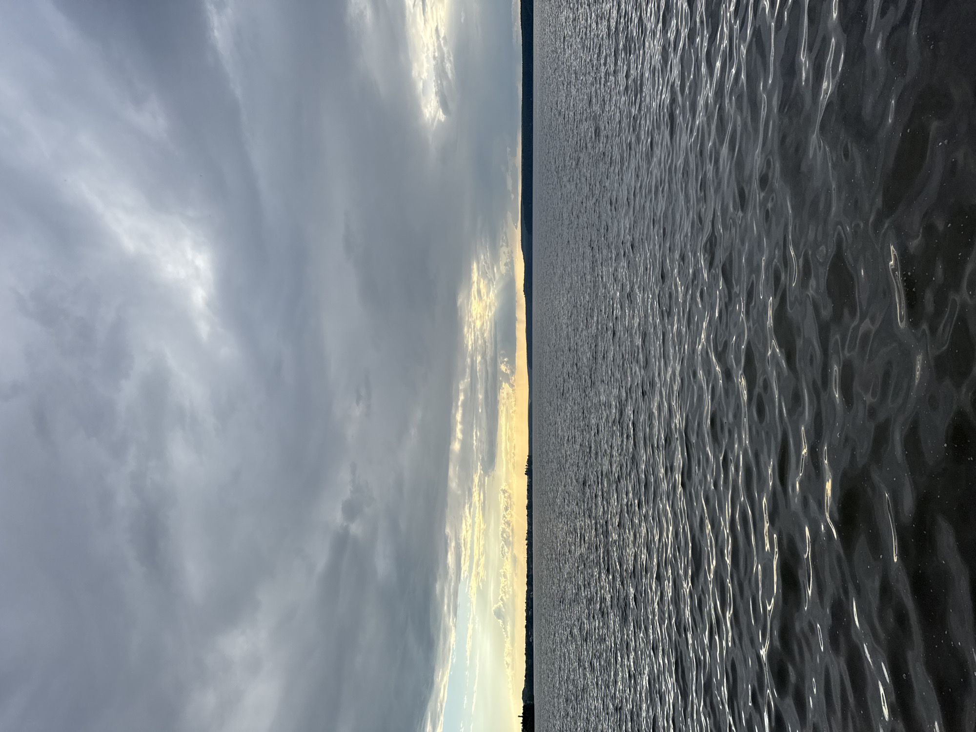 The image depicts a wide expanse of dark, rippling water, likely a lake or a large river, under a cloudy sky. The horizon is marked by a distant, dark silhouette of land with trees. Shafts of sunlight pierce through breaks in the clouds, illuminating a portion of the sky and casting a warm glow on the distant shoreline. The overall mood is serene and slightly dramatic due to the interplay of light and shadow.
The prompt requests this scene to be located in Votkinsk, Russia. Votkinsk is a city in the Udmurt Republic, situated on the banks of the Kama River, a major tributary of the Volga. Therefore, this scene could be a view of the Kama River or one of its associated reservoirs near Votkinsk. There are no people or specific man-made objects clearly identifiable to indicate the exact location within the city. The focus is on the natural landscape of water and sky.