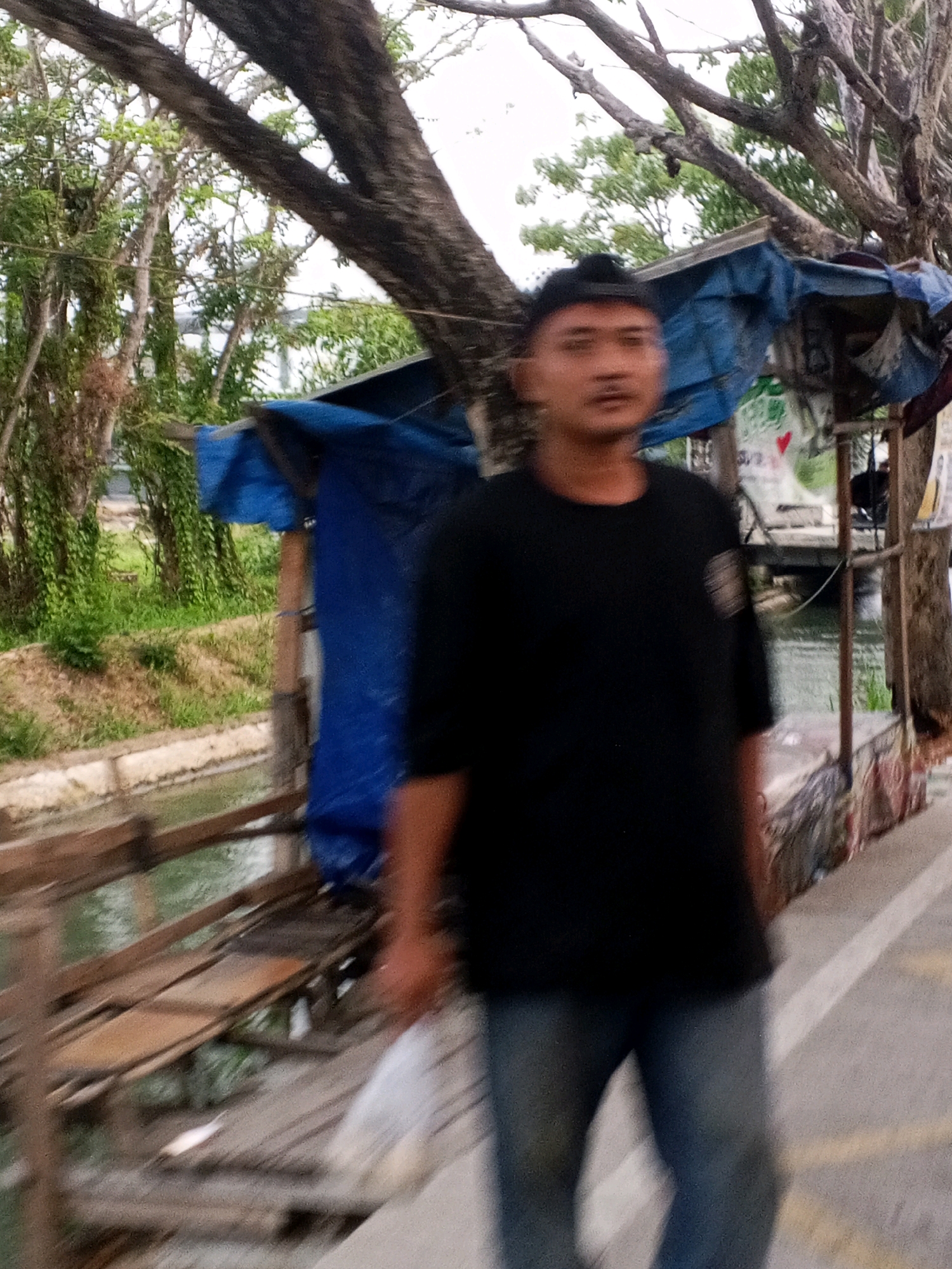 The image shows a blurred, medium-shot of a young man in a black t-shirt and blue jeans standing on a pathway next to a body of water. He is facing the camera with a neutral expression. Behind him, there is a wooden structure covered with blue tarpaulin, and beyond that, lush green vegetation and trees. A wooden bridge or walkway is visible spanning the water. The overall setting appears to be an outdoor, possibly semi-rural area. The image is dated 2023-03-08.